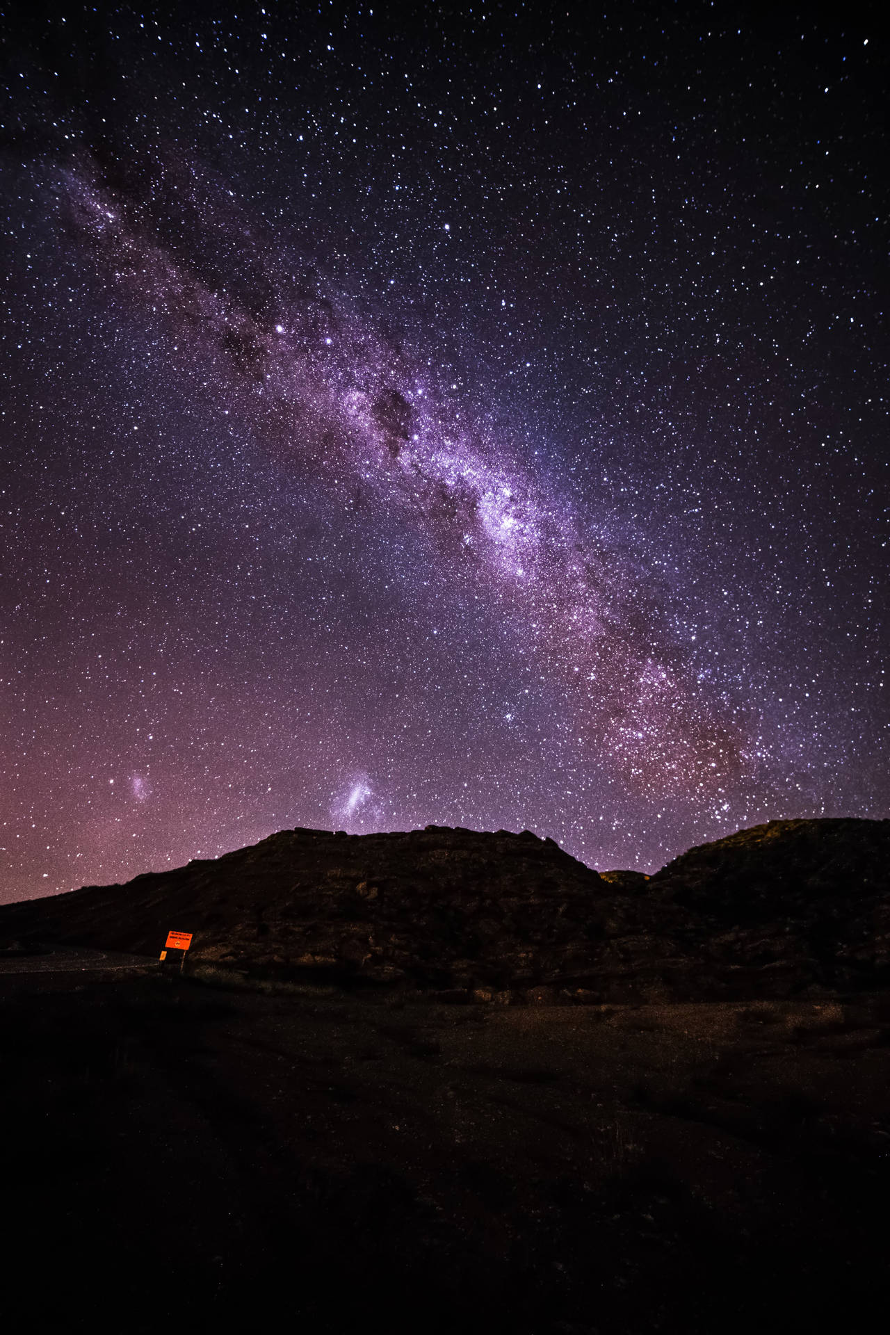 一條紫色銀河系在繁星點點的夜空中延伸，越過黑暗的山丘，上面有一個發光的標誌。