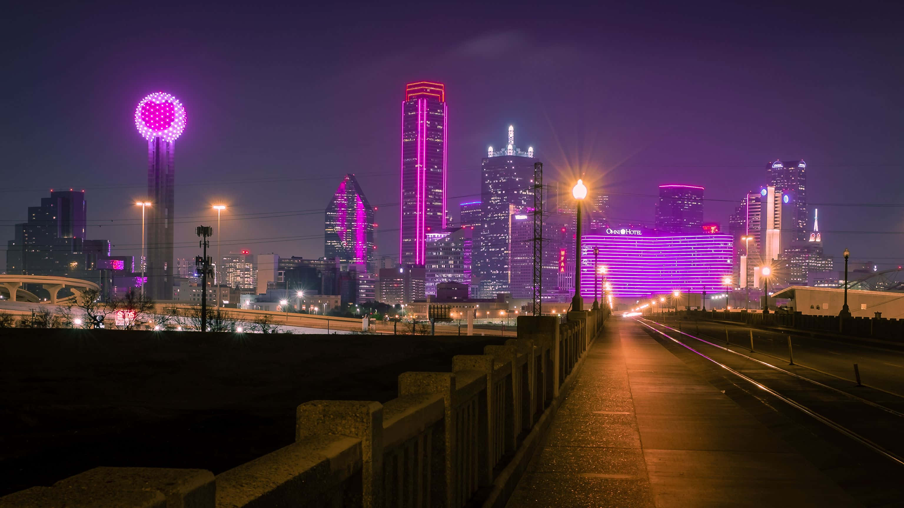 紫色燈光下的達拉斯縣重聚塔背景 壁紙 — 充滿活力的夜間城市景觀，摩天大樓在紫色燈光下閃耀，黑暗的天空下有一個發光的球形塔。