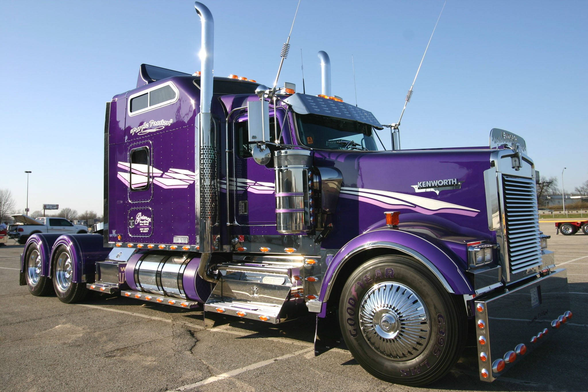 紫色肯沃斯 W900 壁紙 — 一輛閃亮的紫色Kenworth半掛卡車，帶有鍍鉻裝飾和定制貼花，停在陽光明媚的停車場。
