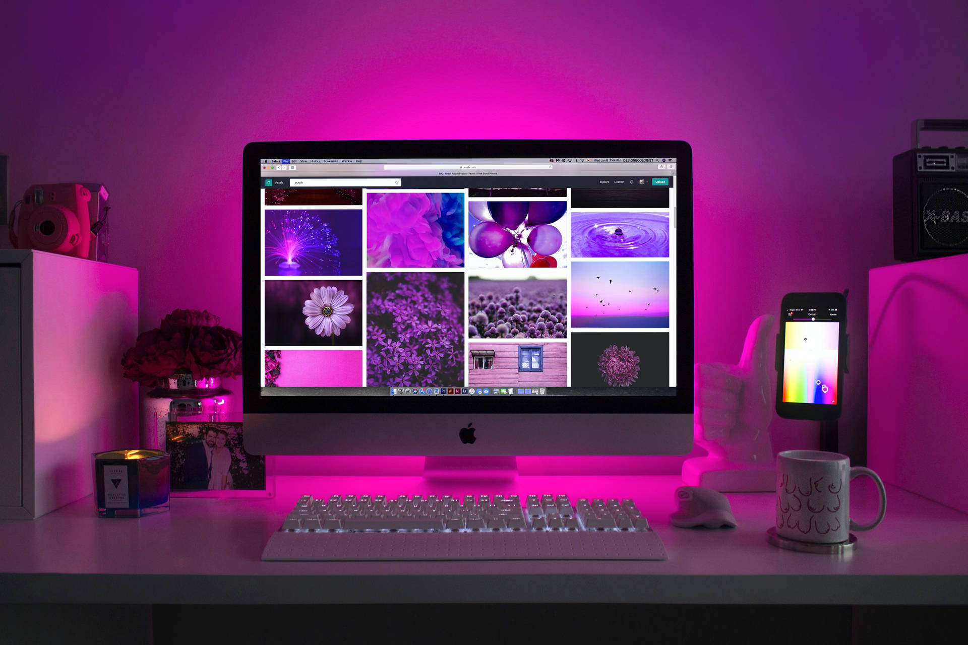 粉紅色照明的書桌，iMac 顯示紫色壁紙畫廊和科技配件。