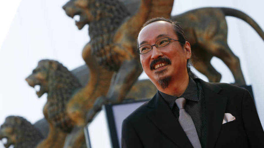 細田守動畫的夢幻場景 壁紙 — 一位微笑的西裝男子在明亮的日光下站在一排青銅獅子雕像前。