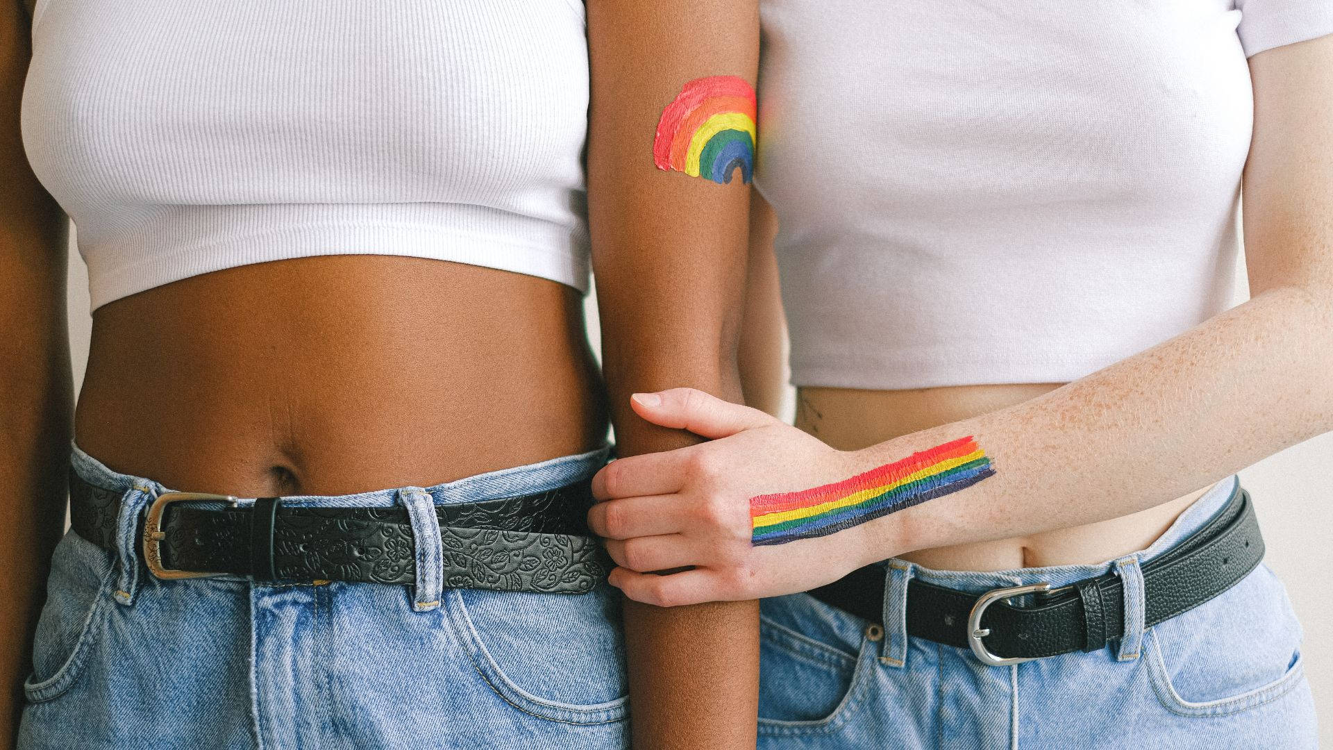 細膩的LGBT彩虹紋身於身體上 壁紙 — 兩個身穿白色短上衣和牛仔褲的人，手臂上裝飾著象徵LGBTQ+驕傲的彩虹臨時紋身。