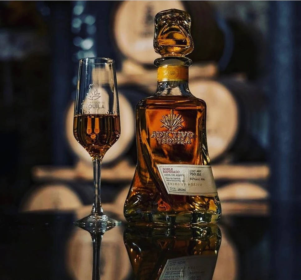 絕美 Adictivo Tequileros 龍舌蘭酒瓶 壁紙 — 一瓶Adictivo Tequila和一個裝滿的玻璃杯在溫暖燈光下閃爍，背景是木桶。