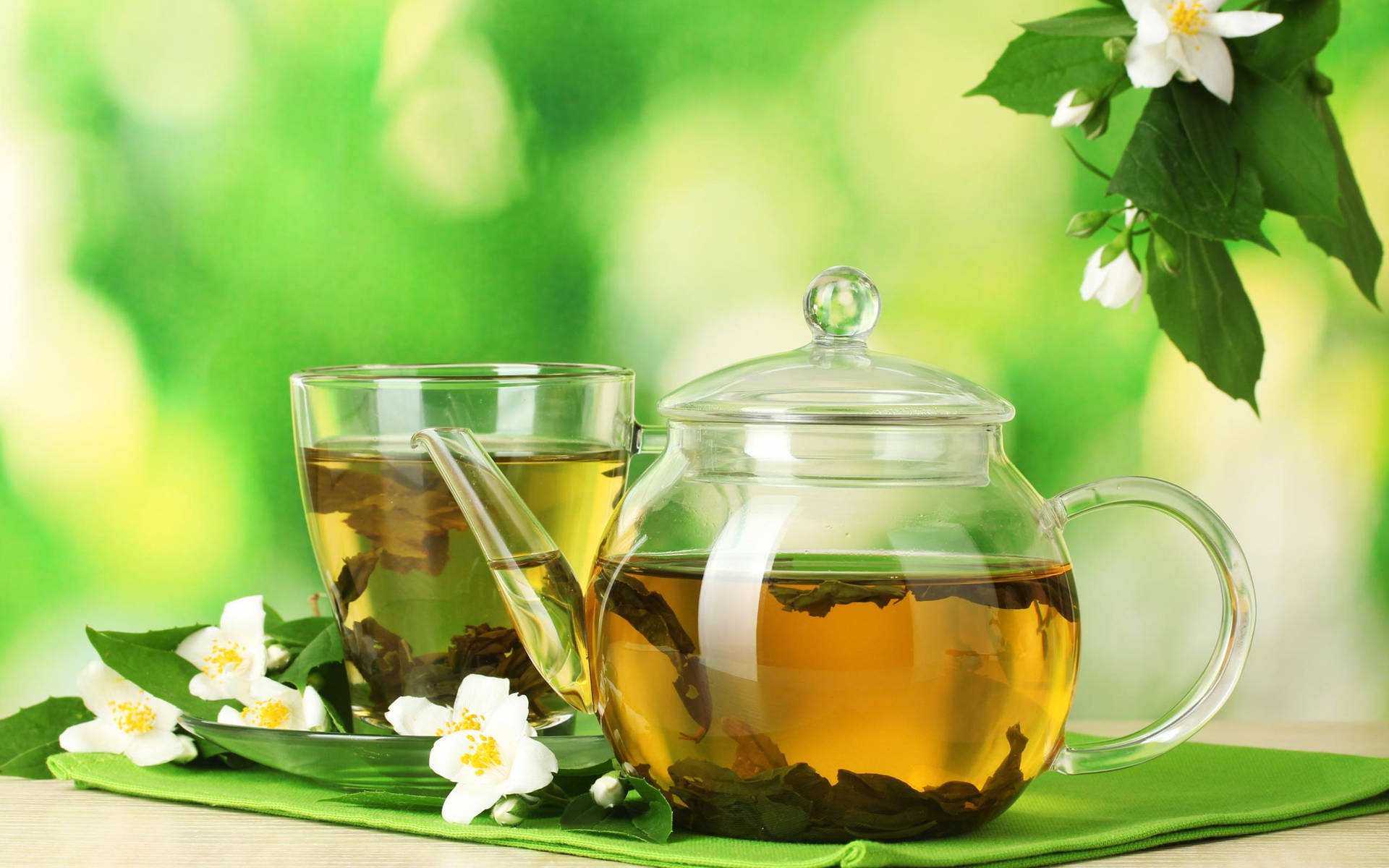 玻璃茶壺和茉莉花茶杯，配上新鮮花朵和葉子，置於綠色布上。