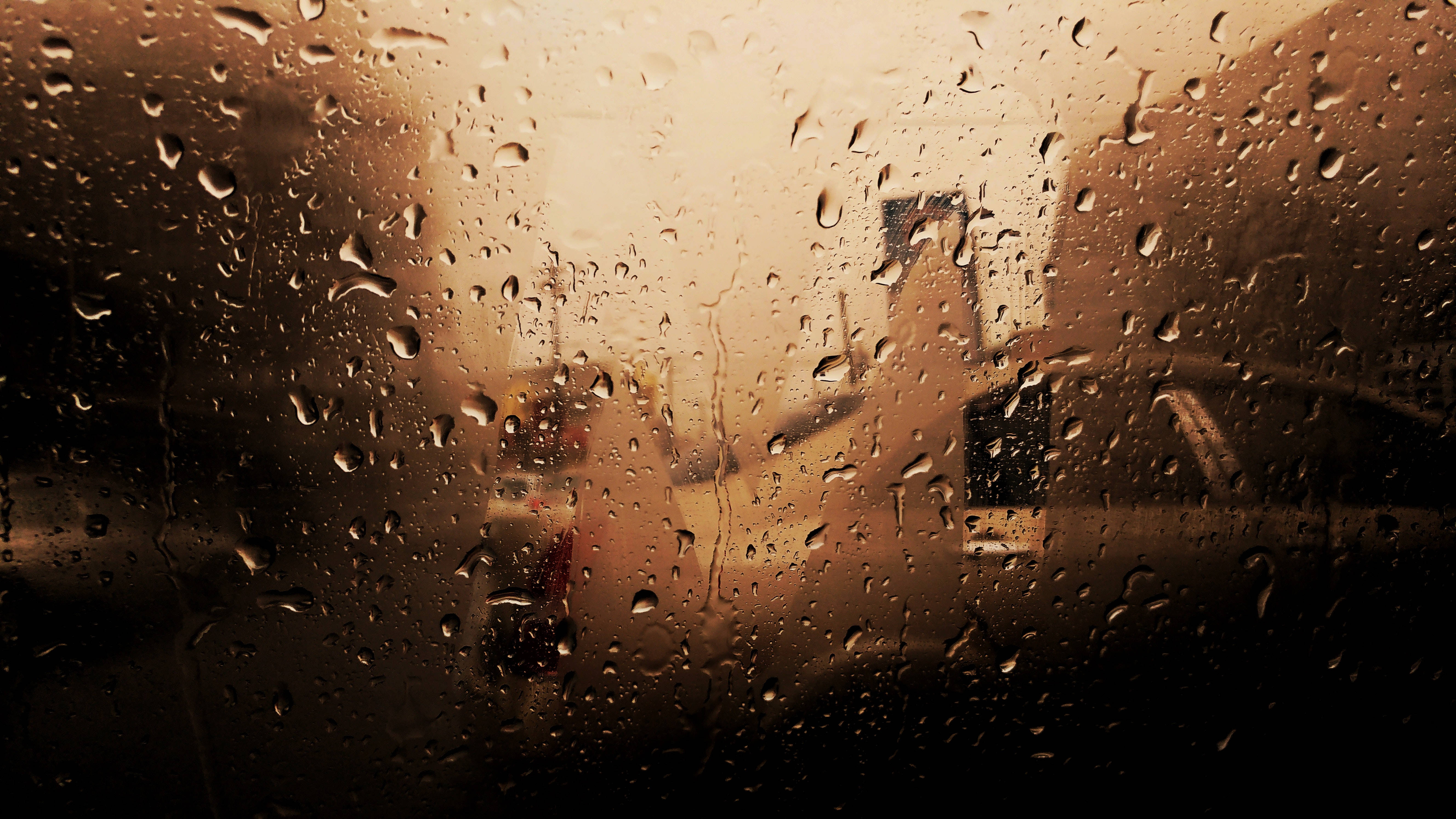 絢爛日落灑落瑙魯海岸 壁紙 — 雨滴劃過窗戶，顯露出黃昏時分模糊的金色城市景觀。