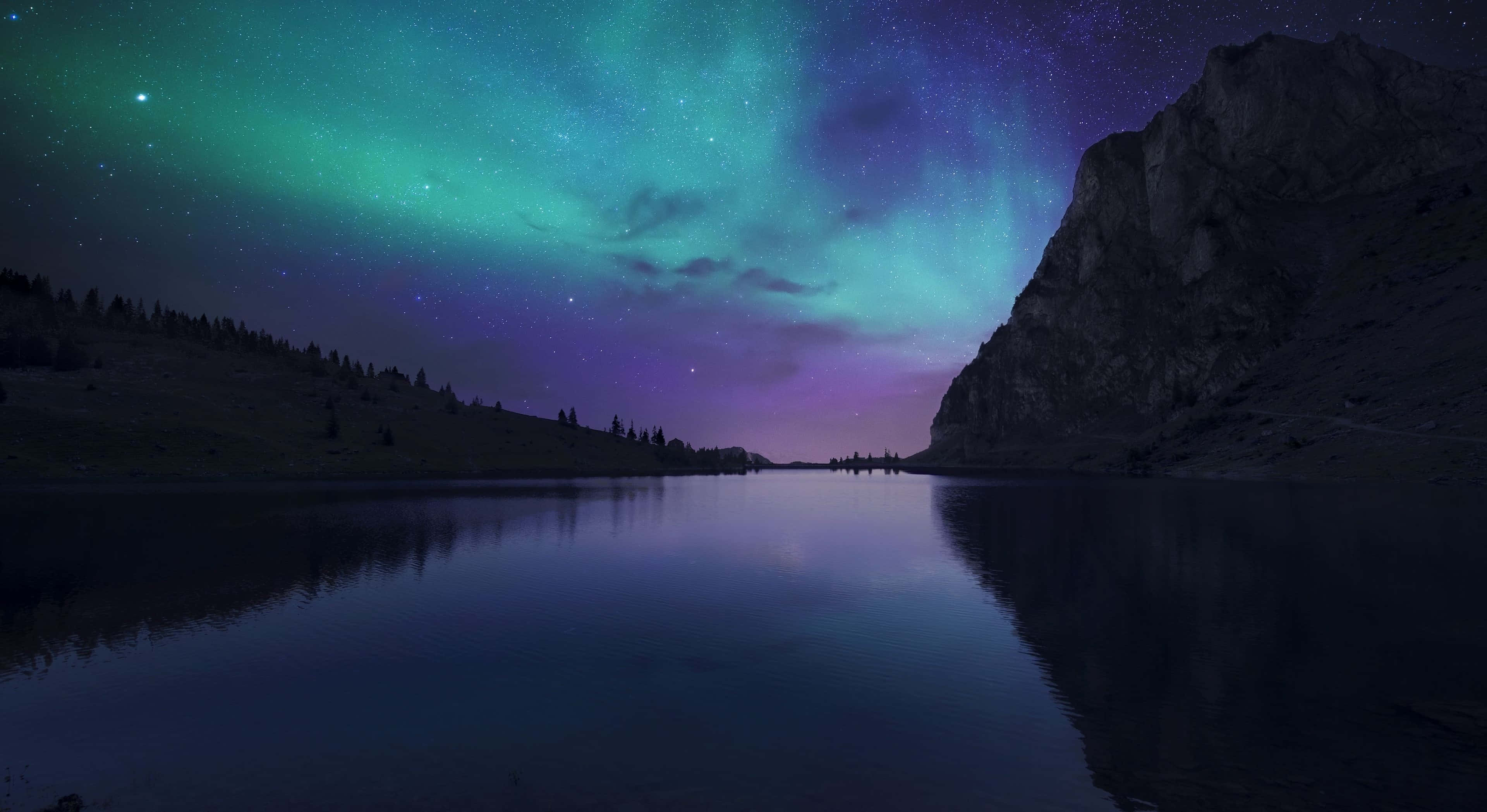 絢麗極光籠罩森林的迷人夜景 壁紙 — 北極光在充滿活力的綠色和紫色中舞動，越過黑暗山脈環繞的平靜湖泊上方的星空夜空。