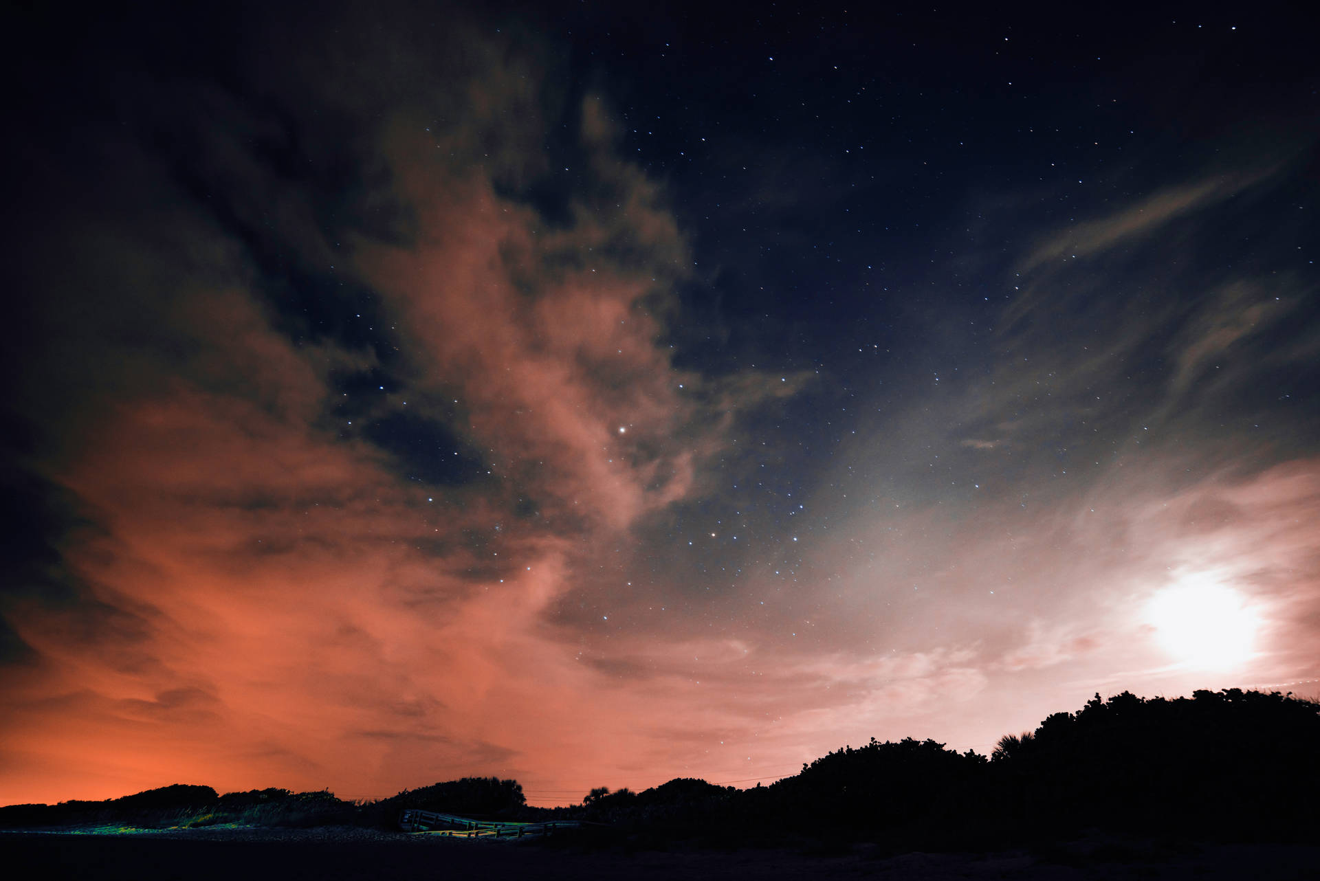 繁星點點的夜空，橙雲和月亮，在黑暗的山丘上方。