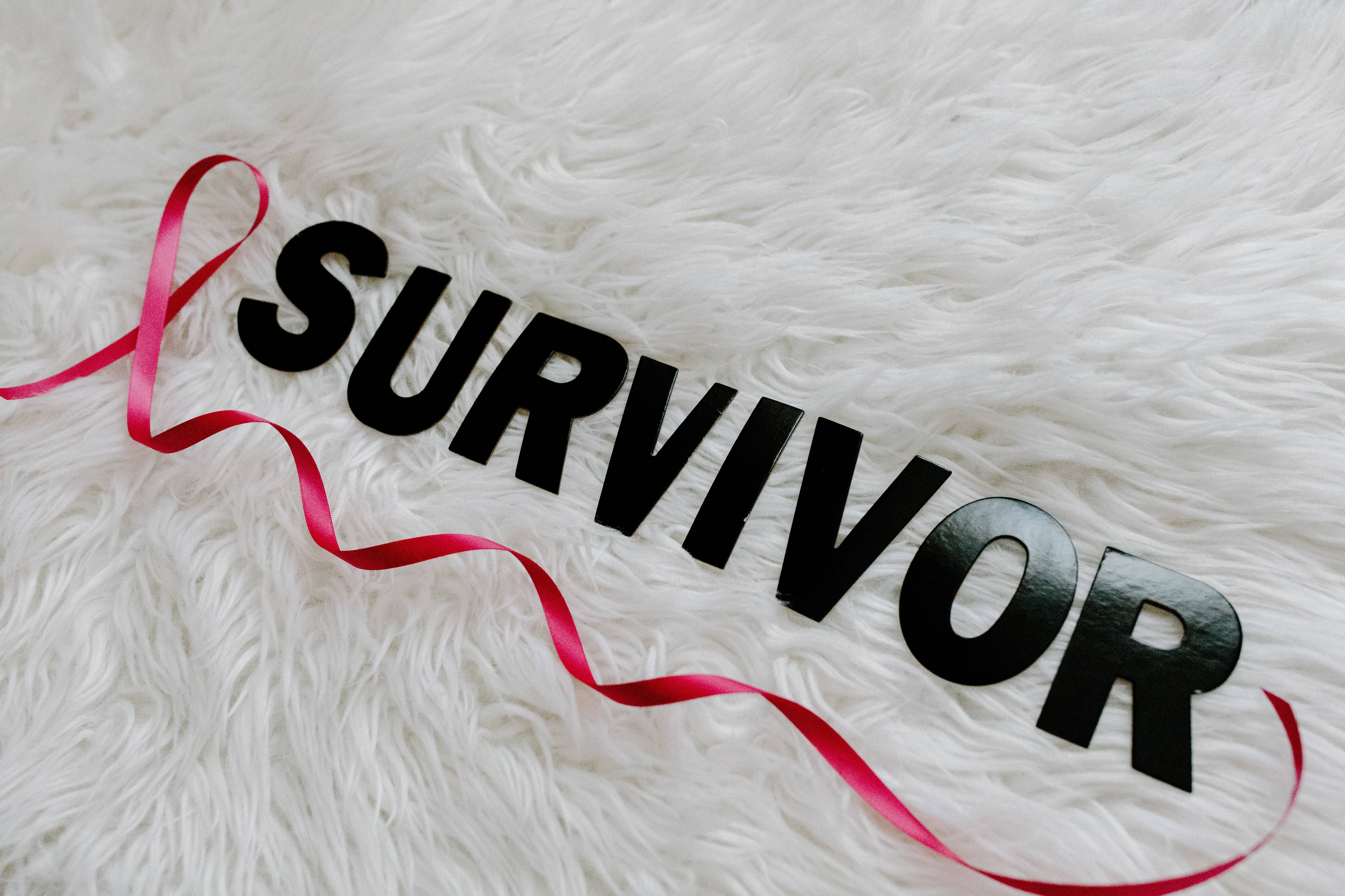 絲帶與生存者 - 乳癌意識 壁紙 — 粗體黑色 'SURVIVOR' 字母和一條紅色絲帶放在柔軟的白色絨毛表面上。