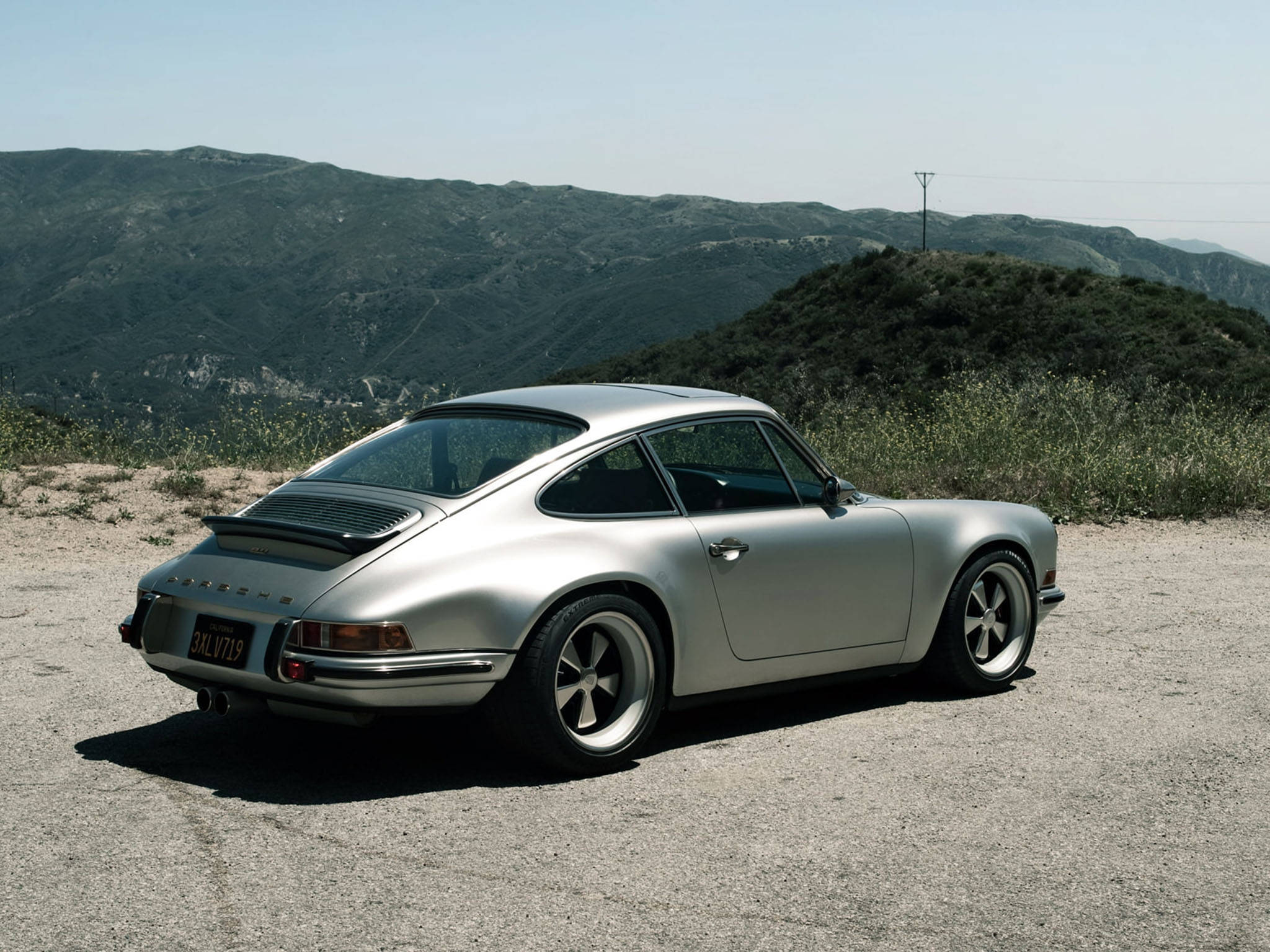 絲絨銀Singer Porsche 壁紙 — 一架時尚的銀色保時捷911經典跑車停泊在塵土飛揚的路邊，背景是連綿起伏的綠色山丘，頭頂是清澈的藍天。