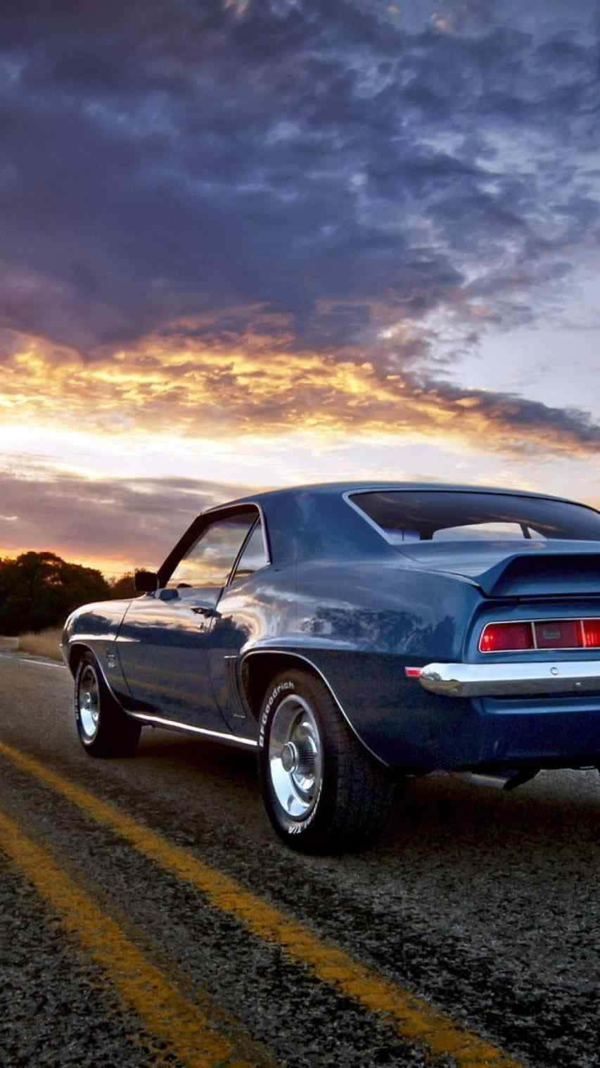 經典 1999 年 Chevrolet Camaro 跑車 iPhone 壁紙 — 一輛經典藍色肌肉車在金色雲彩的戲劇性日落天空下行駛在道路上。