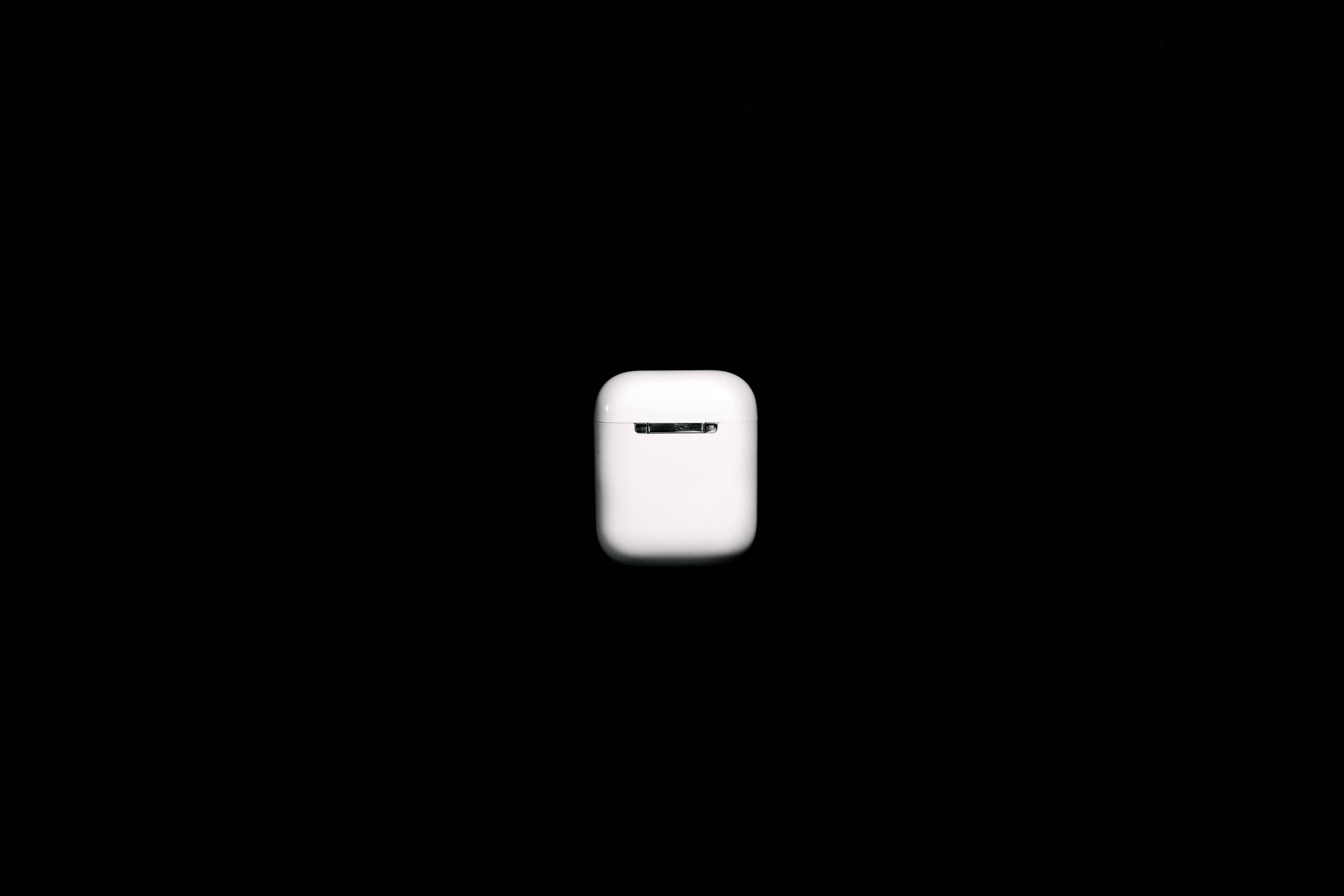經典 Apple AirPods 壁紙 — 一個極簡風格的白色無線耳機充電盒，置於純黑色背景中央，帶有柔和燈光。