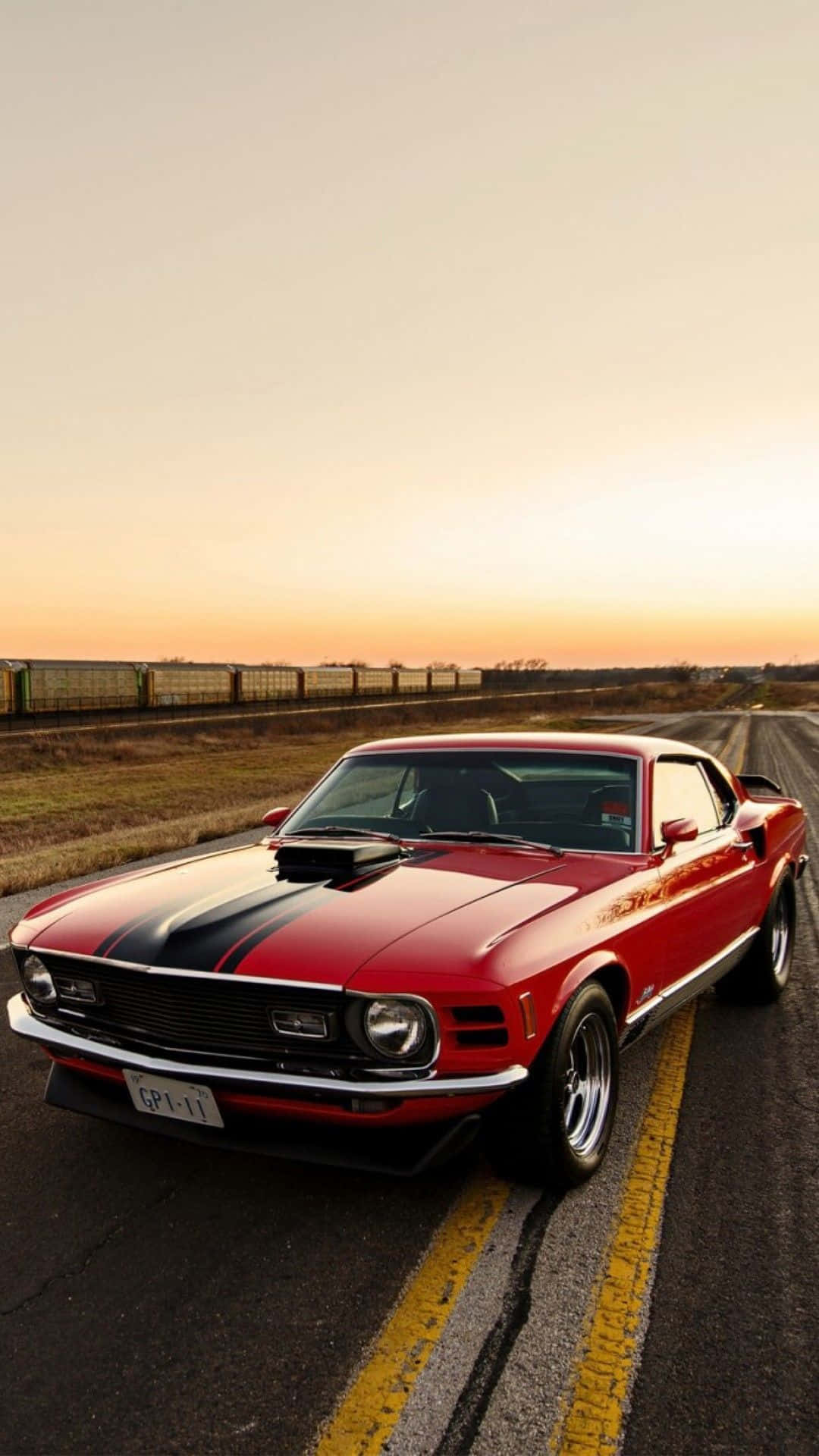 經典 Boss 302 Mustang 跑車 iPhone 壁紙 — 一輛帶有黑色條紋的紅色經典肌肉車在日落時停泊在道路上，背景有火車。