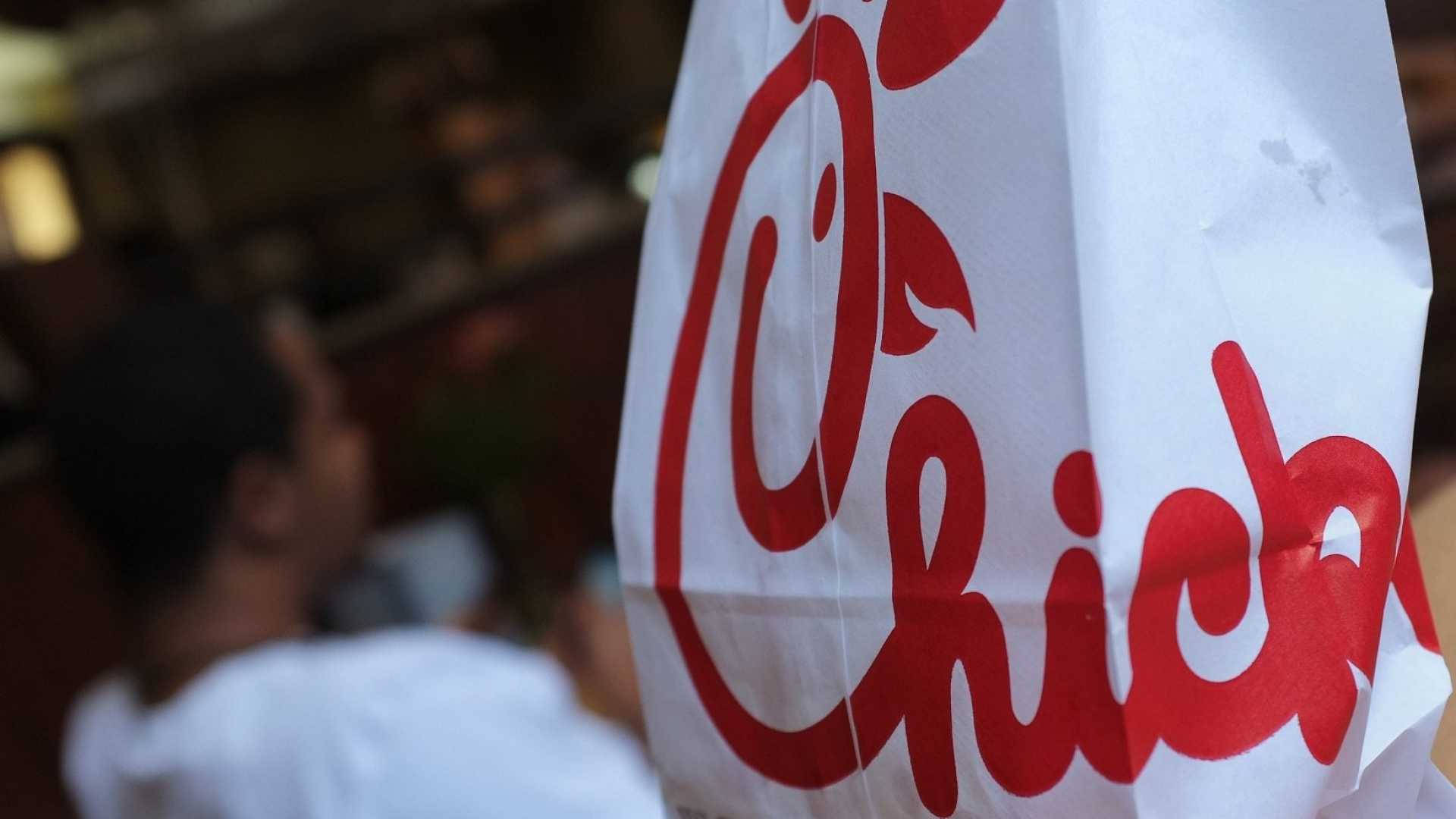 經典 Chick Fil A 白色紙袋 壁紙 — 一個白色Chick-fil-A紙袋的特寫，前景有粗體紅色標誌，背景有模糊的人影。