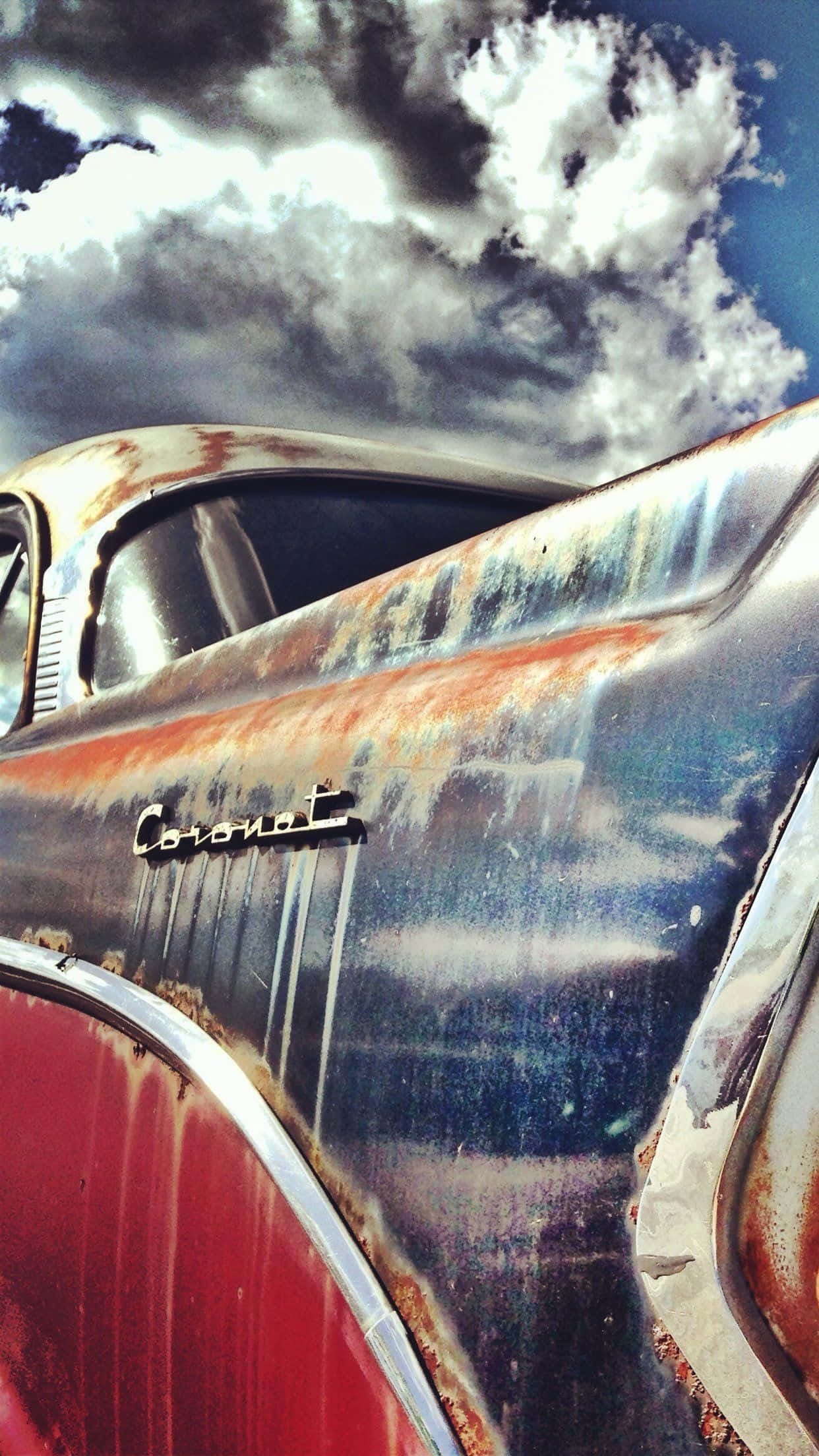 經典 Dodge Coronet 車型 iPhone 壁紙 — 一輛生鏽的復古車，藍色和紅色油漆剝落，在戲劇性的多雲天空下。