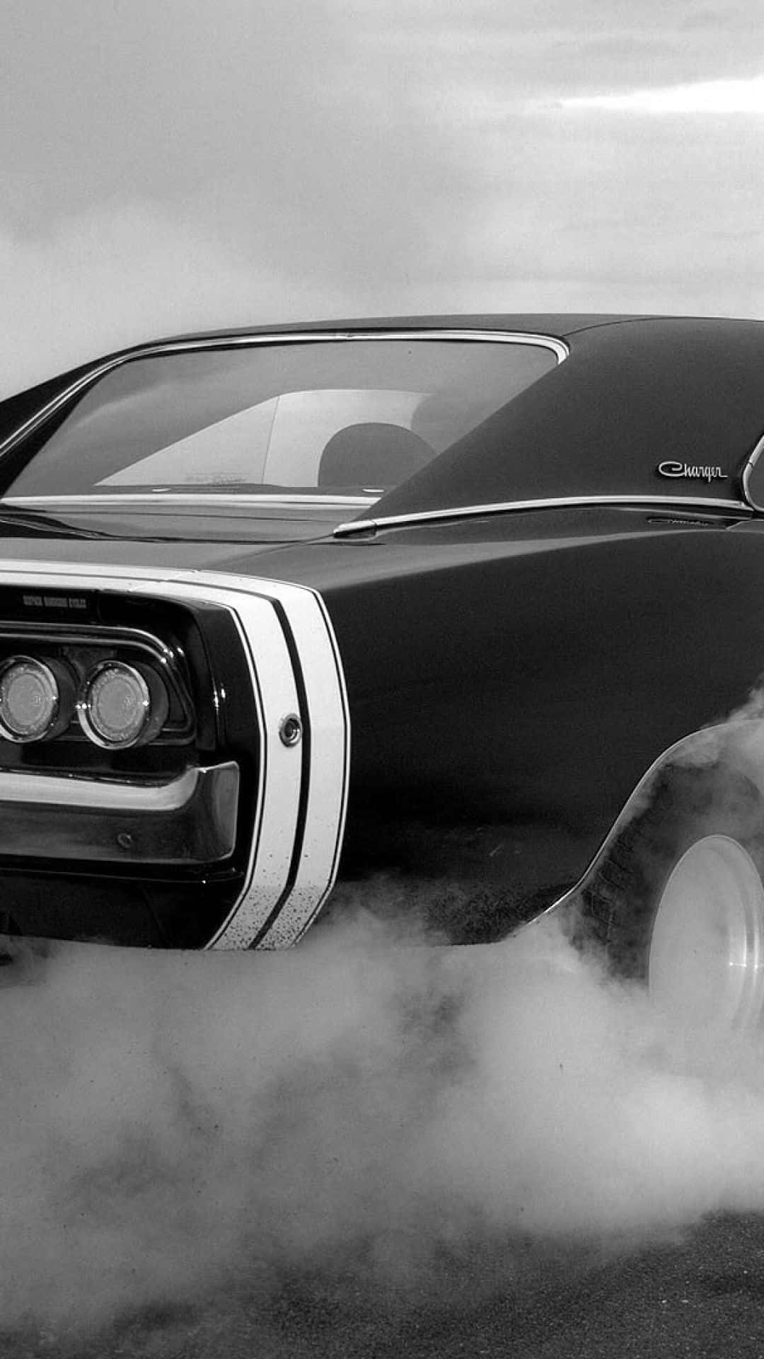 經典 Dodge 跑車 iPhone 壁紙 — 一輛黑色經典肌肉車，帶有鍍鉻細節，後輪冒出白煙，呈現戲劇性的黑白畫面。