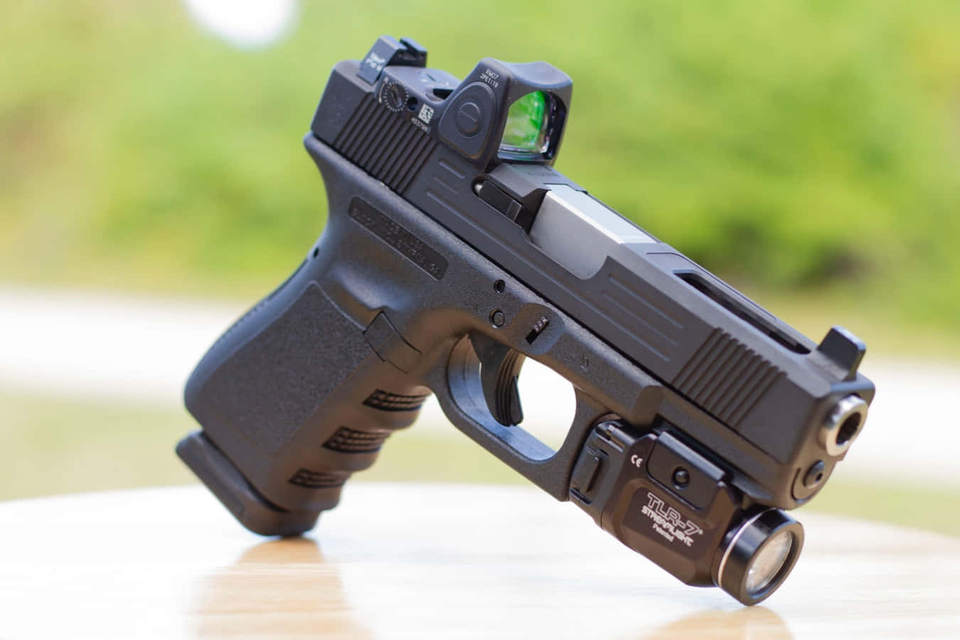經典 Glock 19 手槍 壁紙 — 一把黑色半自動手槍配備紅點瞄準器和戰術燈，放置在木質表面上，背景為模糊的綠色。