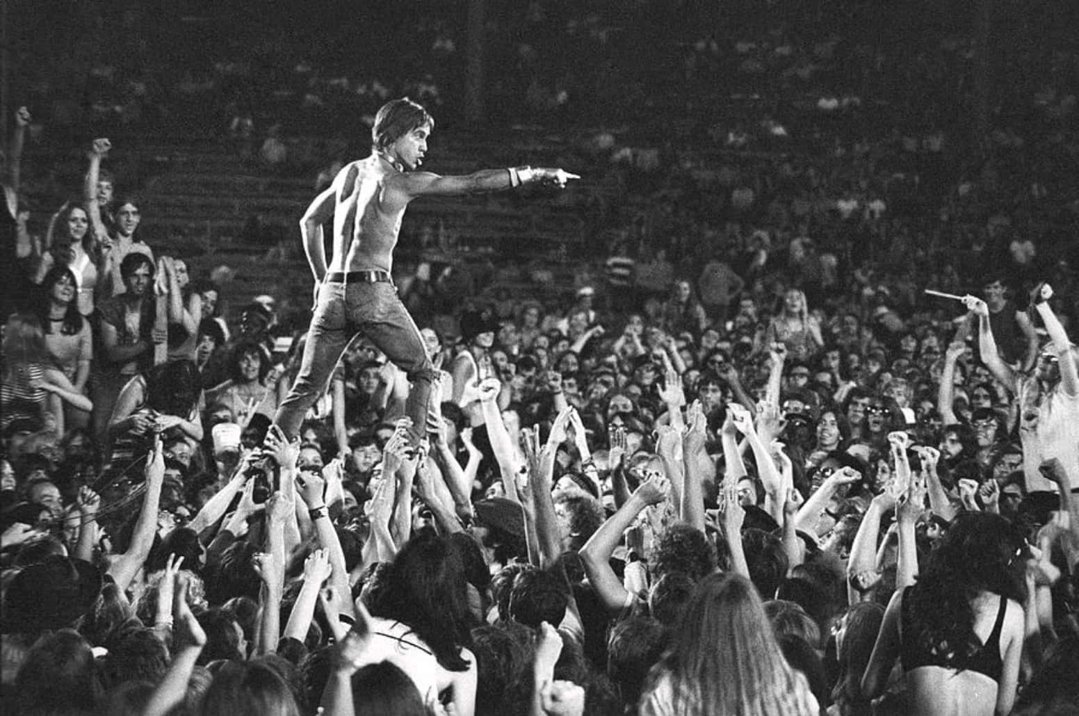 經典 Iggy Pop 演唱會照片 壁紙 — 一位赤裸上身的表演者站在夜燈下擠滿人的演唱會中舉起的手海之上。