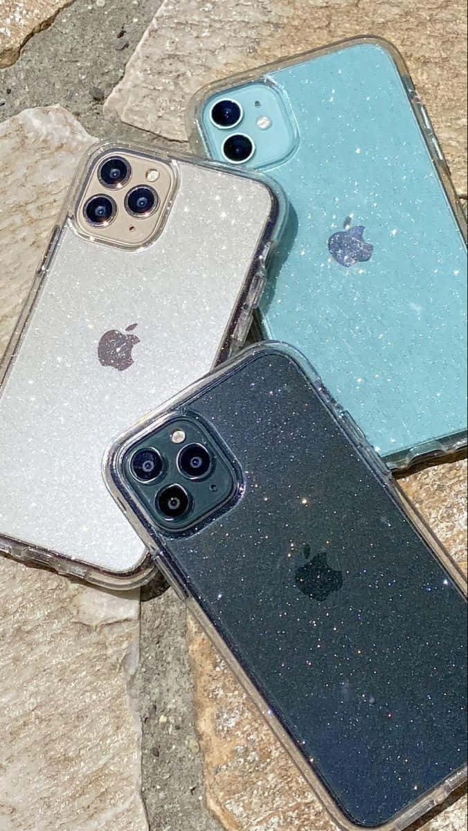 「經典 iPhone 12」 壁紙 — 三個閃粉手機殼，分別為銀色、藍綠色和黑色，在明亮的陽光下放置於粗糙的石頭上。