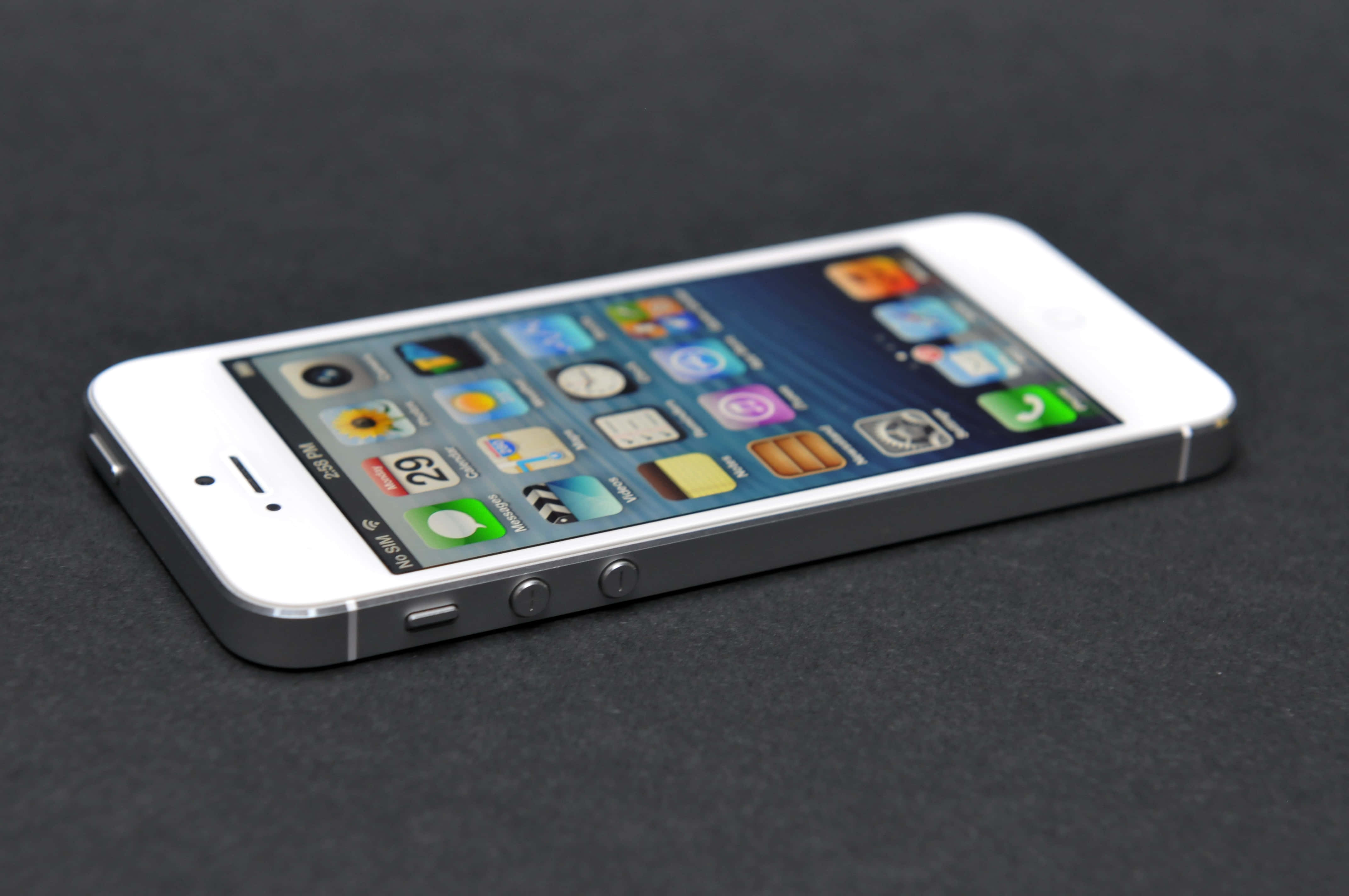 經典 iPhone 的現代演進 壁紙 — 一部白色 iPhone 5s 傾斜放置在深色紋理表面上，其色彩繽紛的應用程式圖標在柔和燈光下發光。