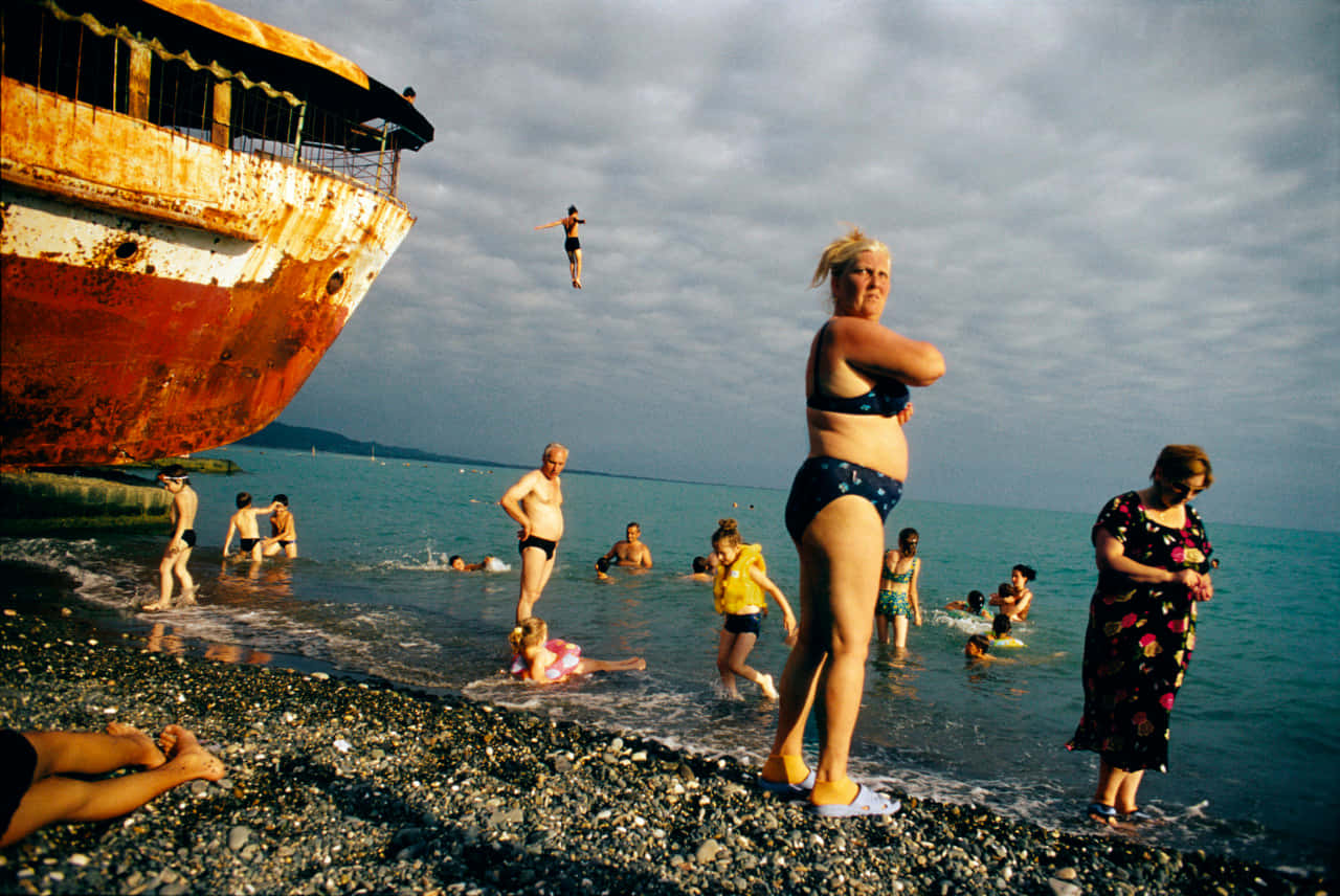 經典 Jonas Bendiksen 巨作 壁紙 — 人們在陰天時享受著生鏽沉船旁邊的鵝卵石海灘，一名游泳者跳入海中。
