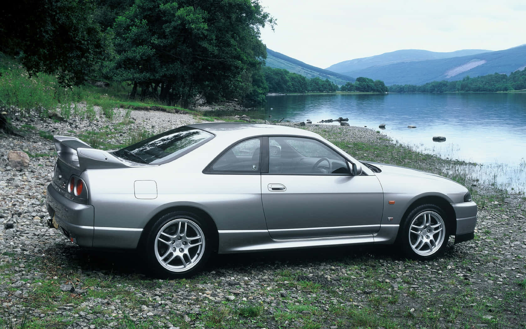 經典 Nissan R33 GTR — 無敵傳奇 壁紙 — 一輛銀色跑車停泊在碎石岸邊，旁邊是一個平靜的湖泊，背景是樹木繁茂的山丘。