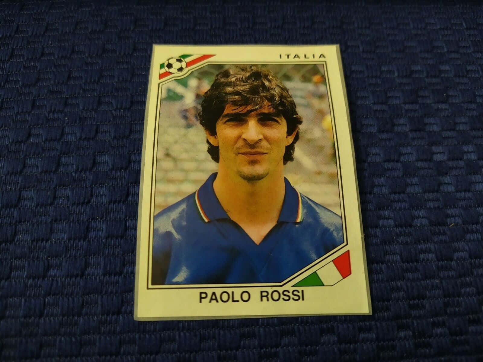 經典 Panini Paolo Rossi 壁紙 — 復古 Paolo Rossi 足球交換卡放置在帶有意大利國旗裝飾的紋理藍色布料上。