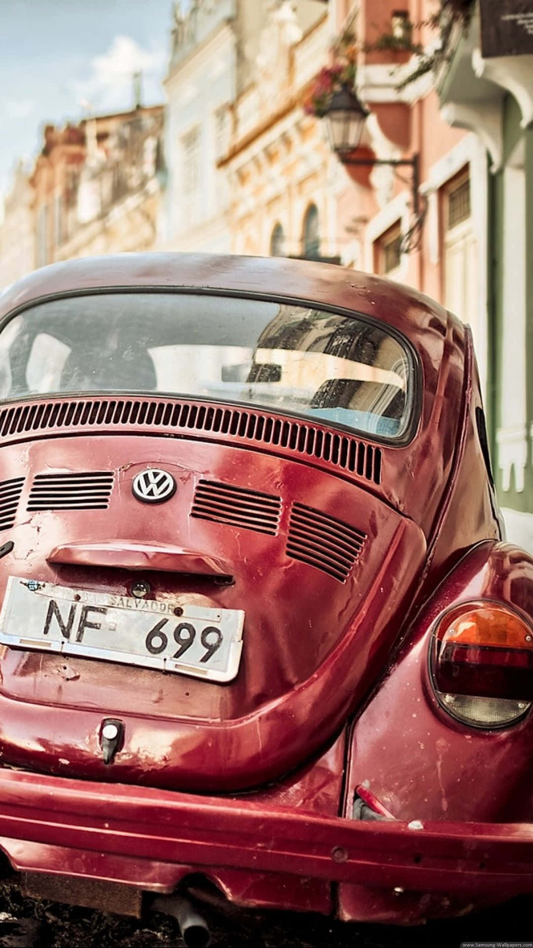 經典 Volkswagen Beetle 跑車 iPhone 壁紙 — 一輛風化的紅色復古福士甲蟲車停泊在陽光明媚的歐洲街道上，背景是粉彩色的建築物。