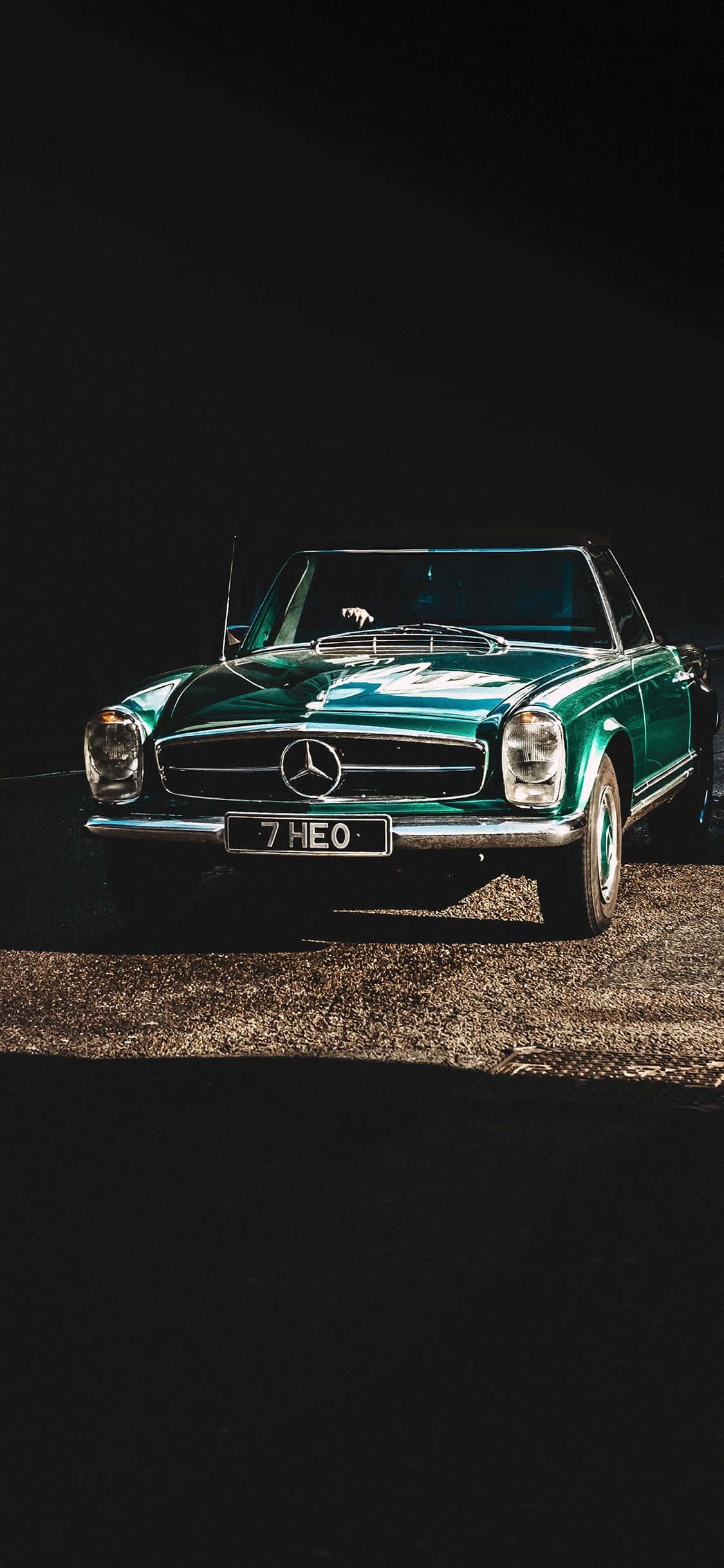 經典 W113 Mercedes-Benz 車型 iPhone 壁紙 — 一輛復古綠色平治SL開篷車在黑色背景的碎石表面上，於戲劇性的聚光燈下閃閃發光。