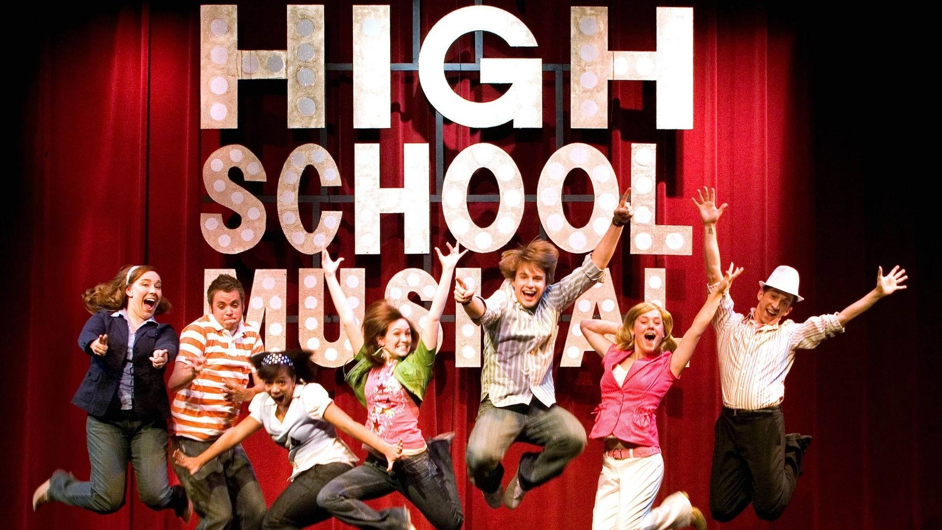 經典《歌舞青春》 壁紙 — 七位快樂的演員在紅色帷幕前，於發光的「HIGH SCHOOL MUSICAL」標誌前充滿活力地跳躍。
