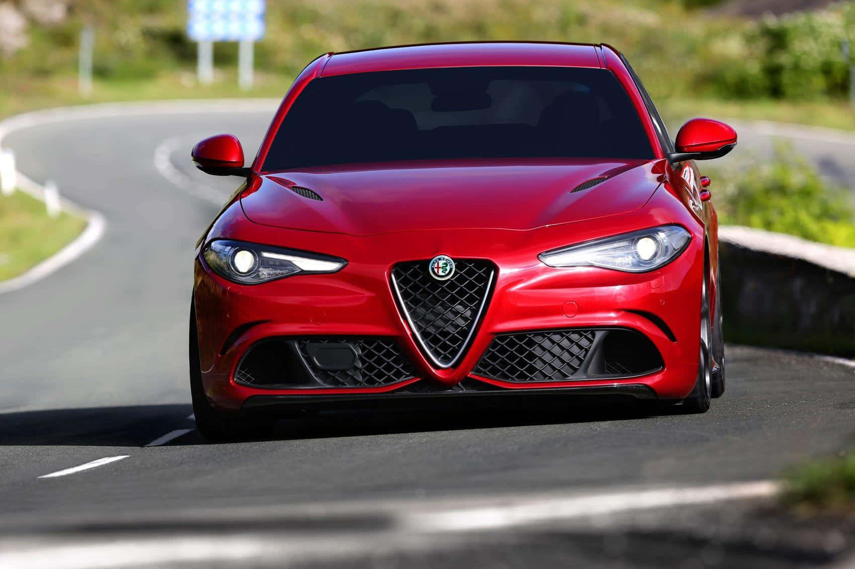 經典Alfa Romeo Giulia 壁紙 — 一輛鮮紅色的愛快羅密歐跑車在明亮的日光下，沿著蜿蜒的瀝青道路飛馳，周圍綠樹成蔭。