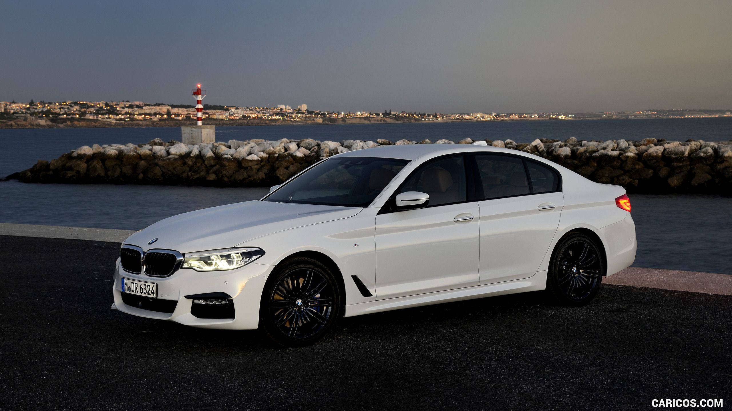 經典BMW M系列跑車 壁紙 — 一架時尚的白色寶馬房車在黃昏時停泊在海濱，背景有燈塔和遠處的小鎮。