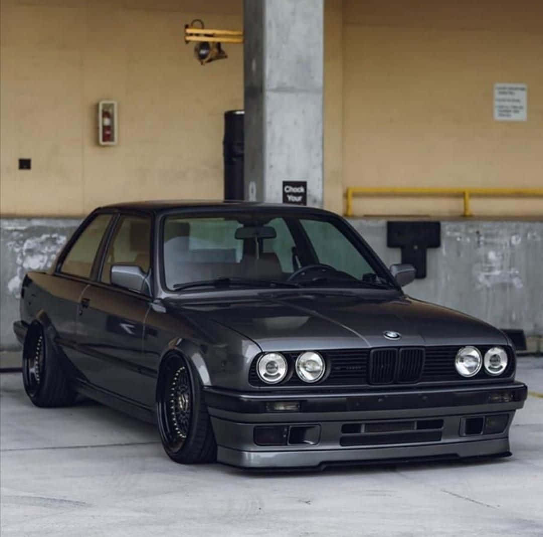 經典BMW車款映襯壯麗自然風光 壁紙 — 一輛啞光黑色的BMW E30雙門轎跑車配備定制輪圈，在混凝土停車場的人造燈光下低趴停放。