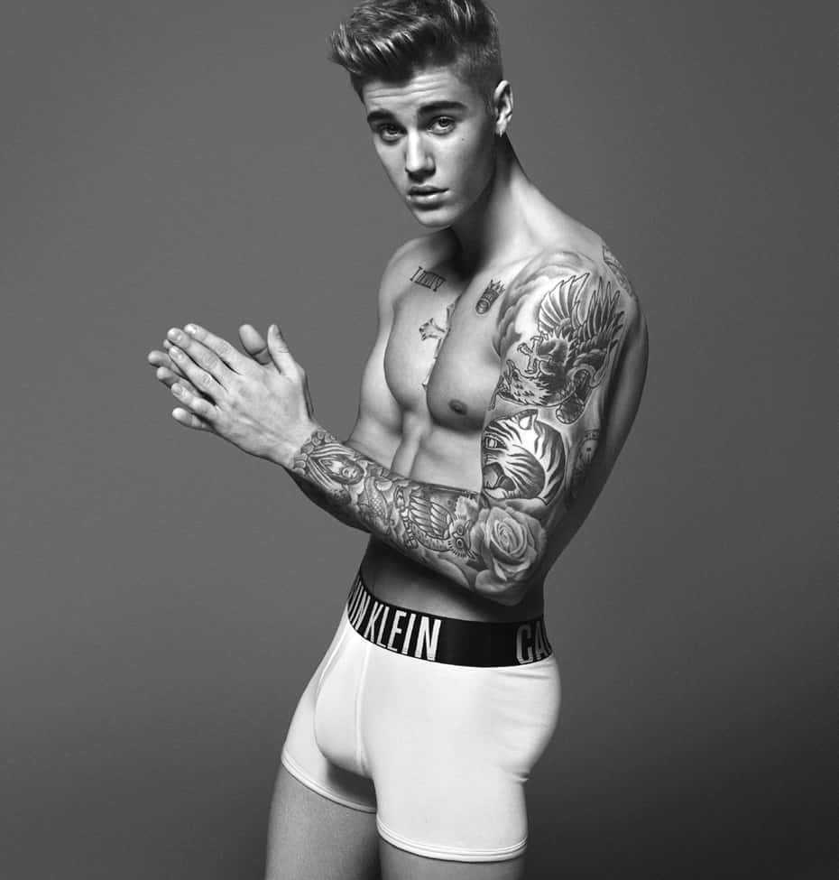 經典Calvin Klein時尚系列 壁紙 — 一位肌肉發達的年輕男子，手臂和胸部有精細的紋身，自信地穿著白色Calvin Klein內褲，在純灰色攝影棚背景前擺姿勢。