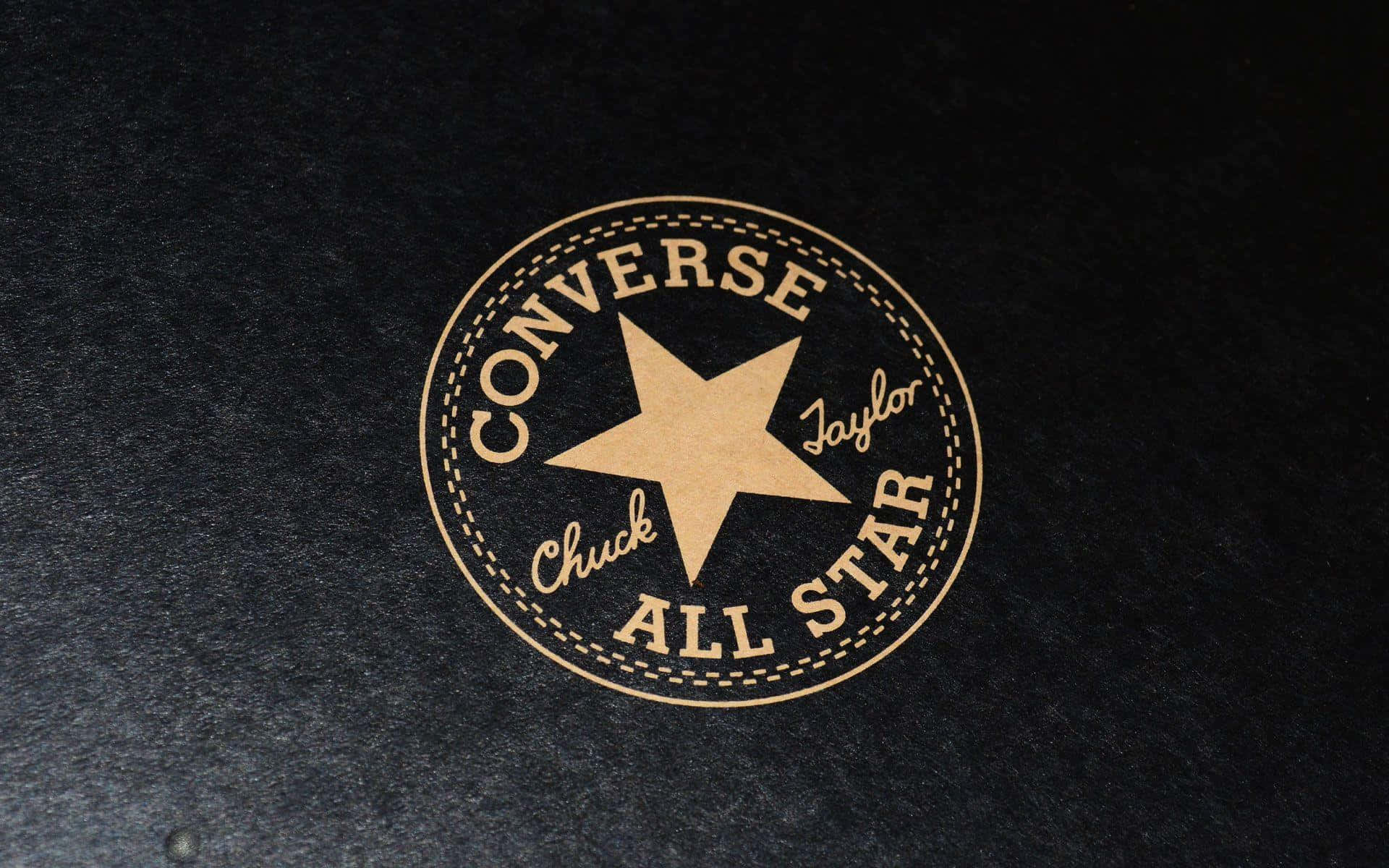 經典Converse All-Star運動鞋堆疊圖 壁紙 — 經典 Converse Chuck Taylor All Star 標誌，以棕褐色呈現於有紋理的黑色布料上。
