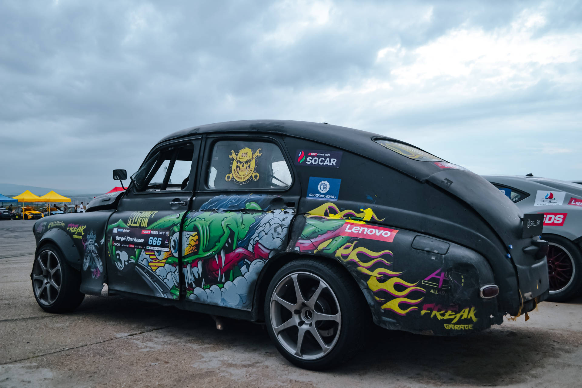 在陰天天空下，賽道上定製塗裝的復古直線加速賽車與塗鴉藝術。