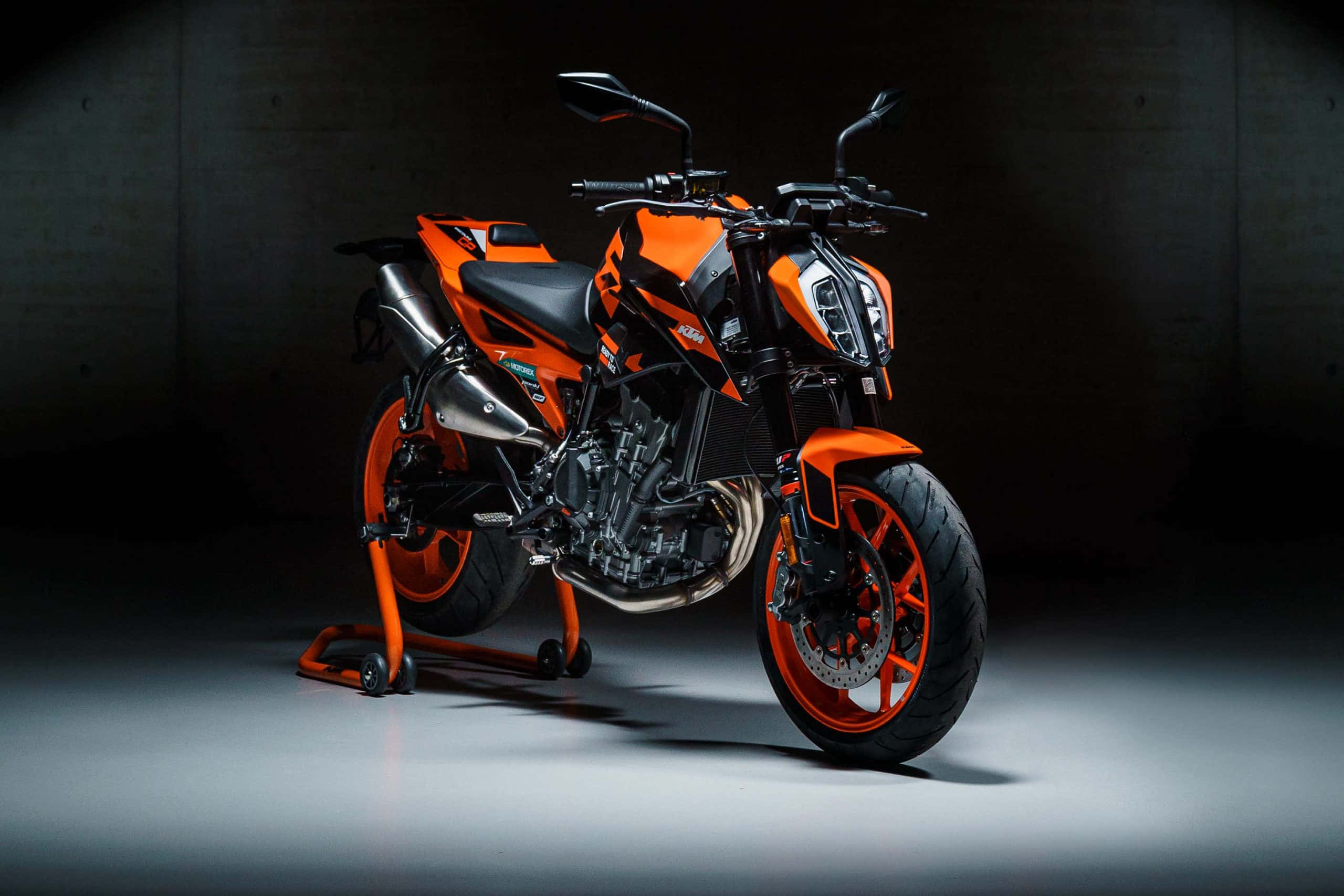 經典KTM Duke 壁紙 — 一架搶眼的橙色和黑色KTM電單車在燈光昏暗的工作室中，在戲劇性的聚光燈下立於支架上。