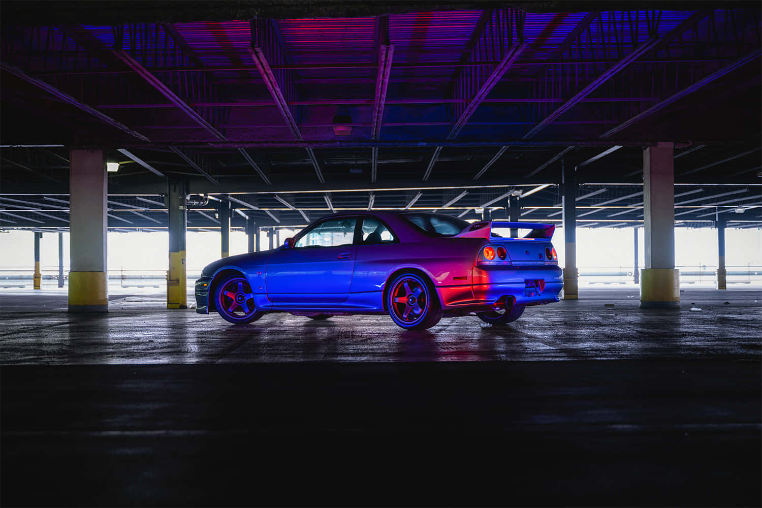 經典Nissan R33 GTR 壁紙 — 一輛藍色跑車在昏暗的地下停車場中，在霓虹紫色和粉紅色燈光下閃耀。