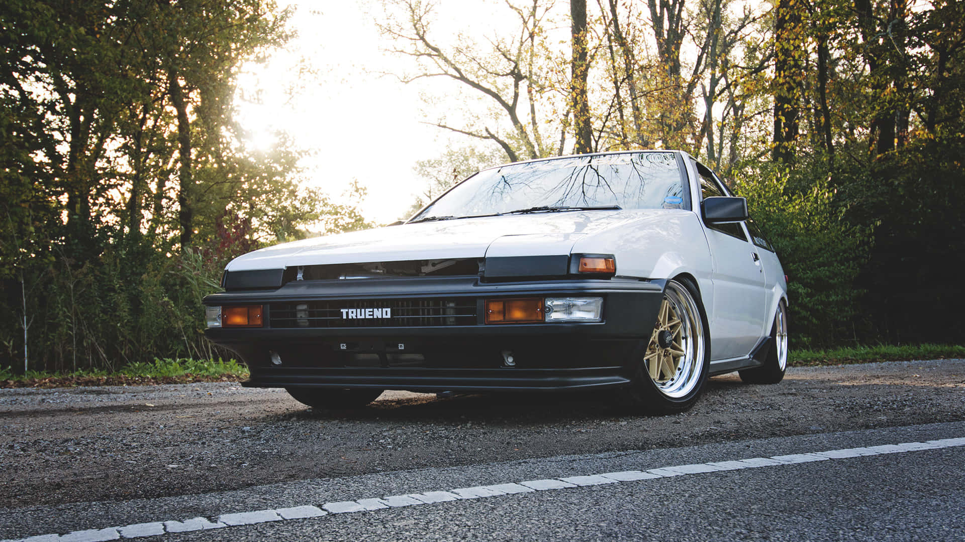 白色豐田 Trueno AE86 配金色輪圈，停泊在日落時分的森林道路上。