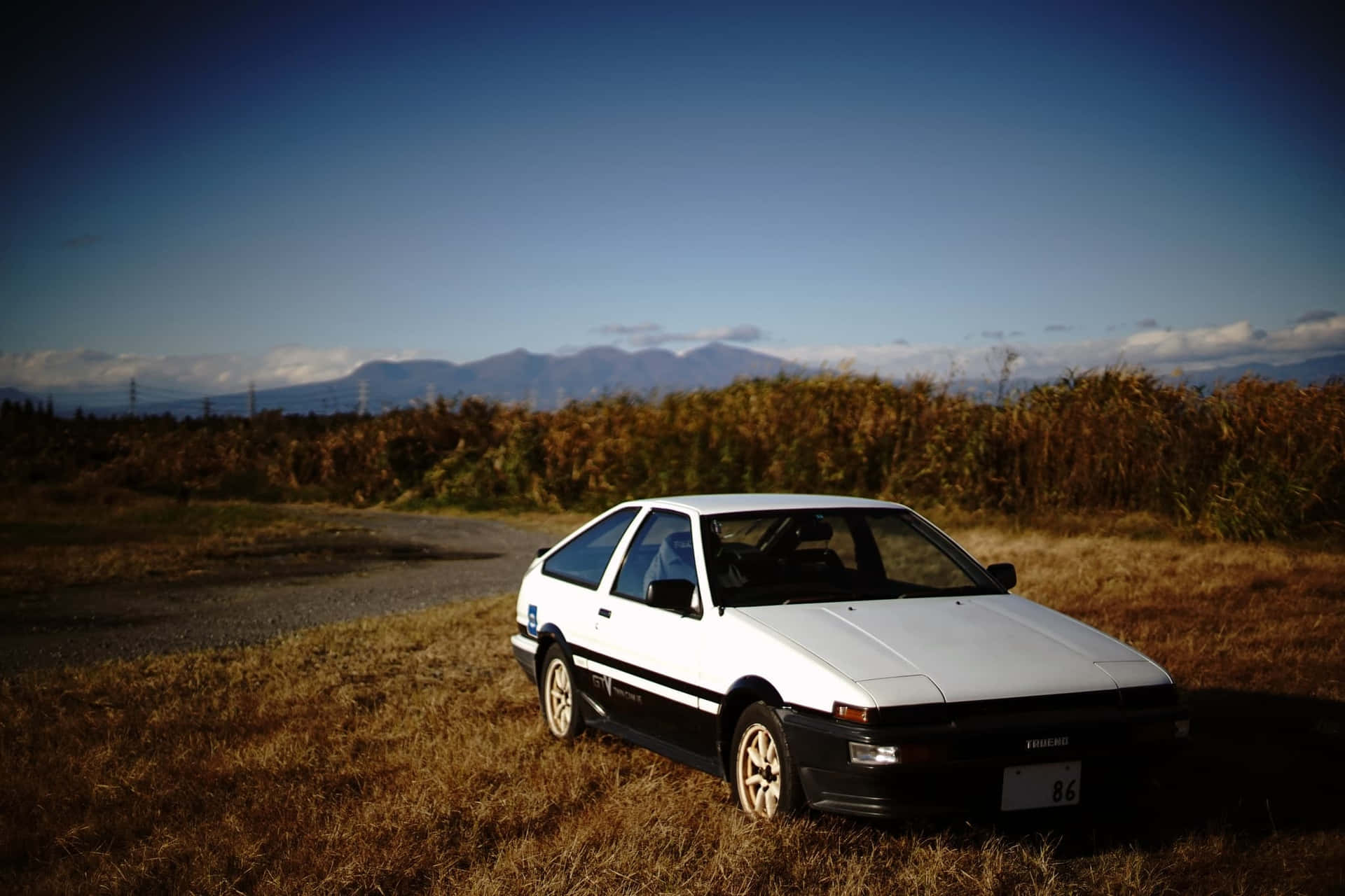 白色AE86跑車停泊在田野中，背景有山。