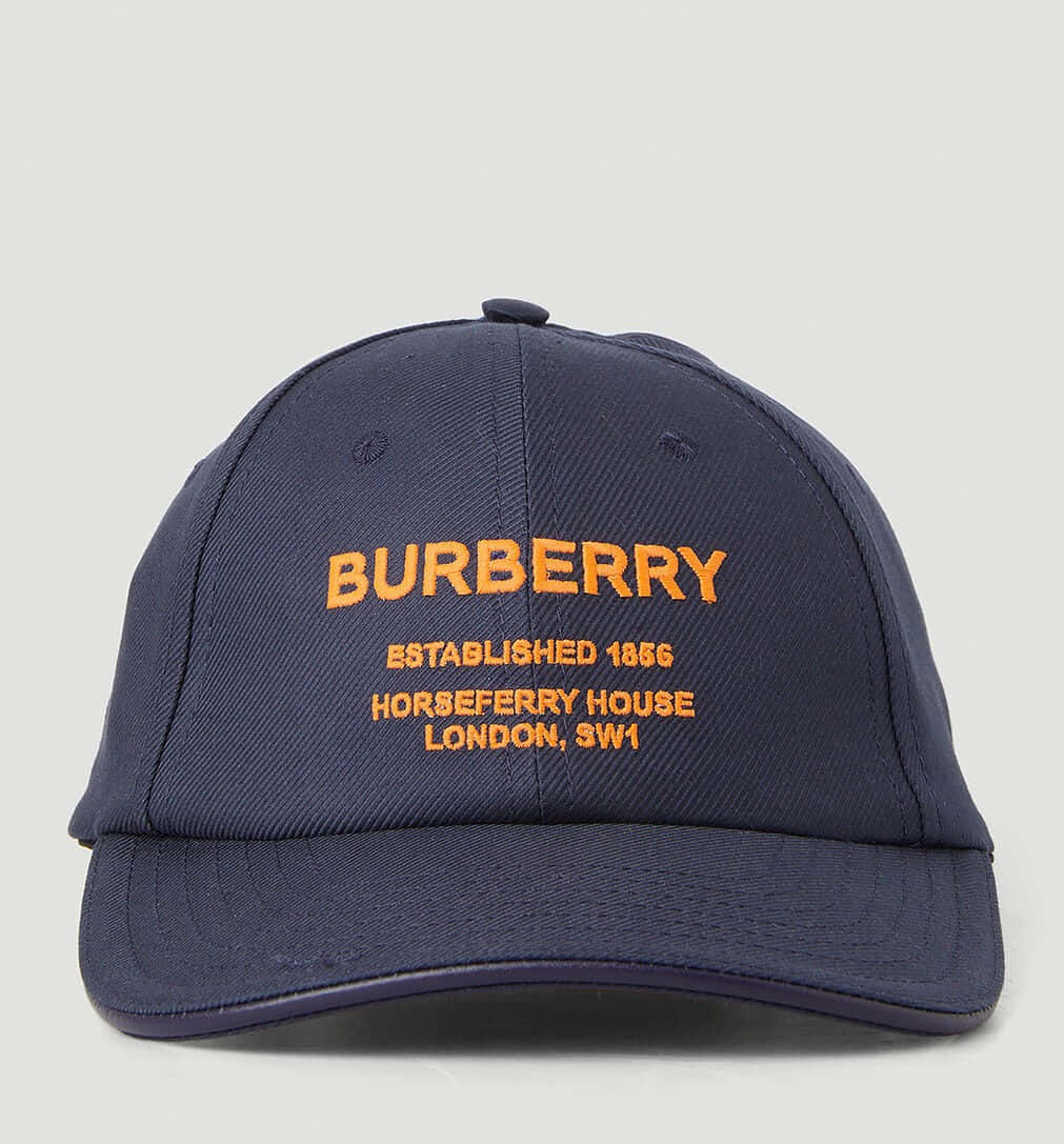 經典傳承：Burberry 的經典設計 壁紙 — 深藍色 Burberry 棒球帽，帶有橙色刺繡文字，詳細說明其傳統和倫敦地址，背景為純色。
