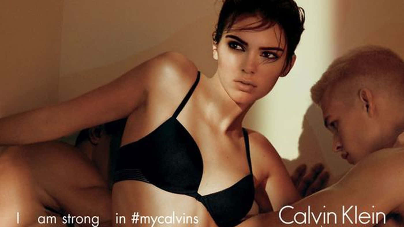 經典優雅的Calvin Klein造型 壁紙 — 一名身穿黑色內衣的女子凝視著鏡頭，旁邊一名光頭男子在溫暖的燈光下低頭看著。