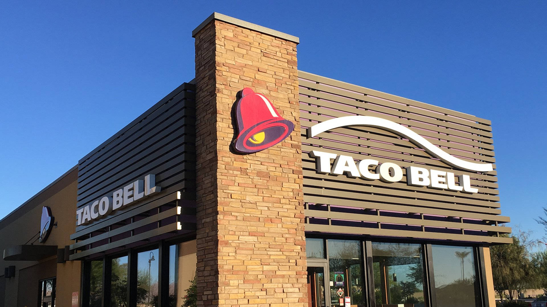 經典塔可鐘餐廳 壁紙 — 鮮紅色的Taco Bell鐘形標誌在磚柱上閃耀，映襯著餐廳現代化外牆上方清澈的藍天。
