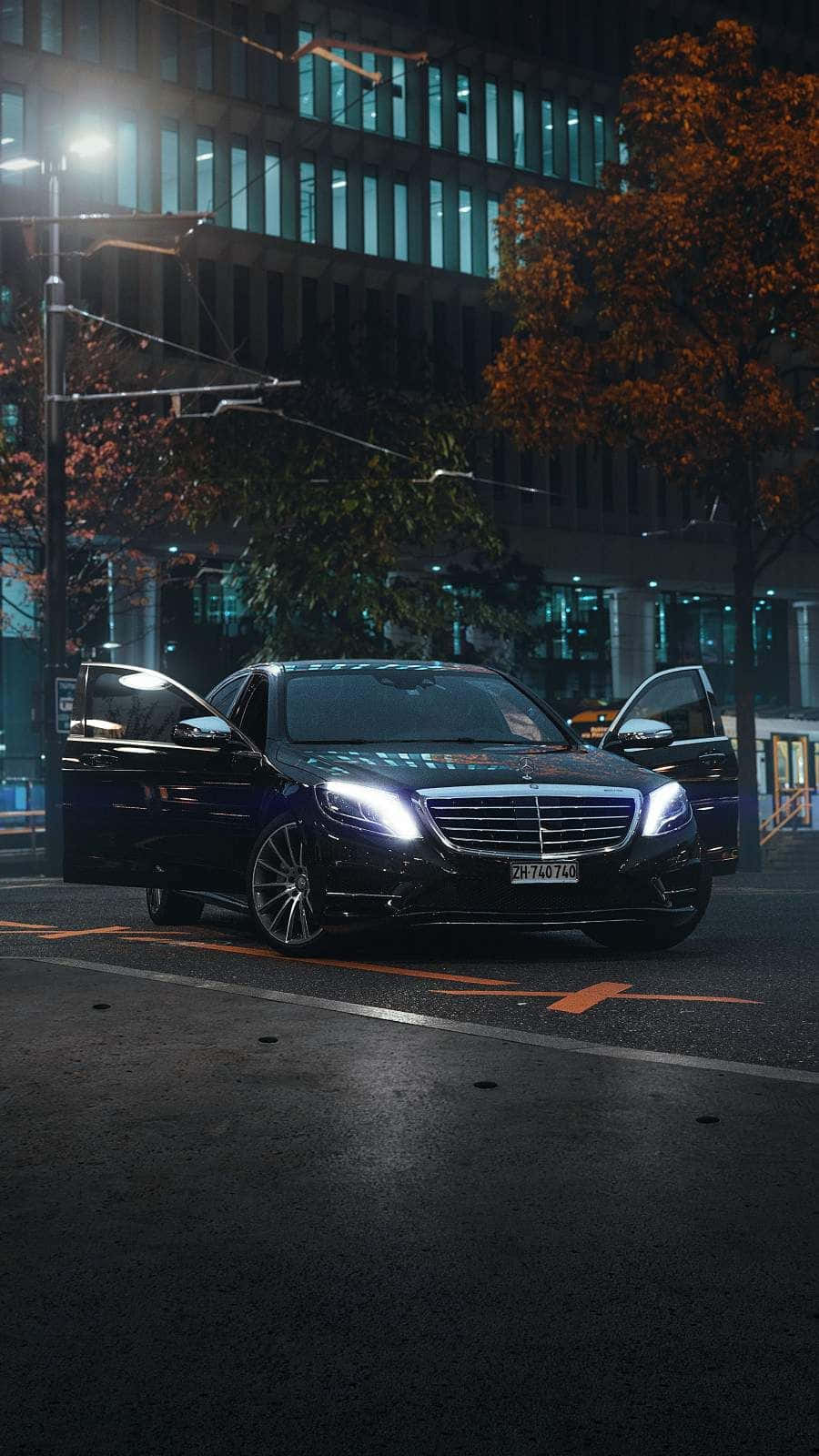 經典奢華：梅賽德斯-奔馳E級 壁紙 — 一輛時尚的黑色梅賽德斯轎車，車頭燈閃爍，停在潮濕的城市街道上，夜晚路燈下，背景有秋樹和亮燈的建築物。