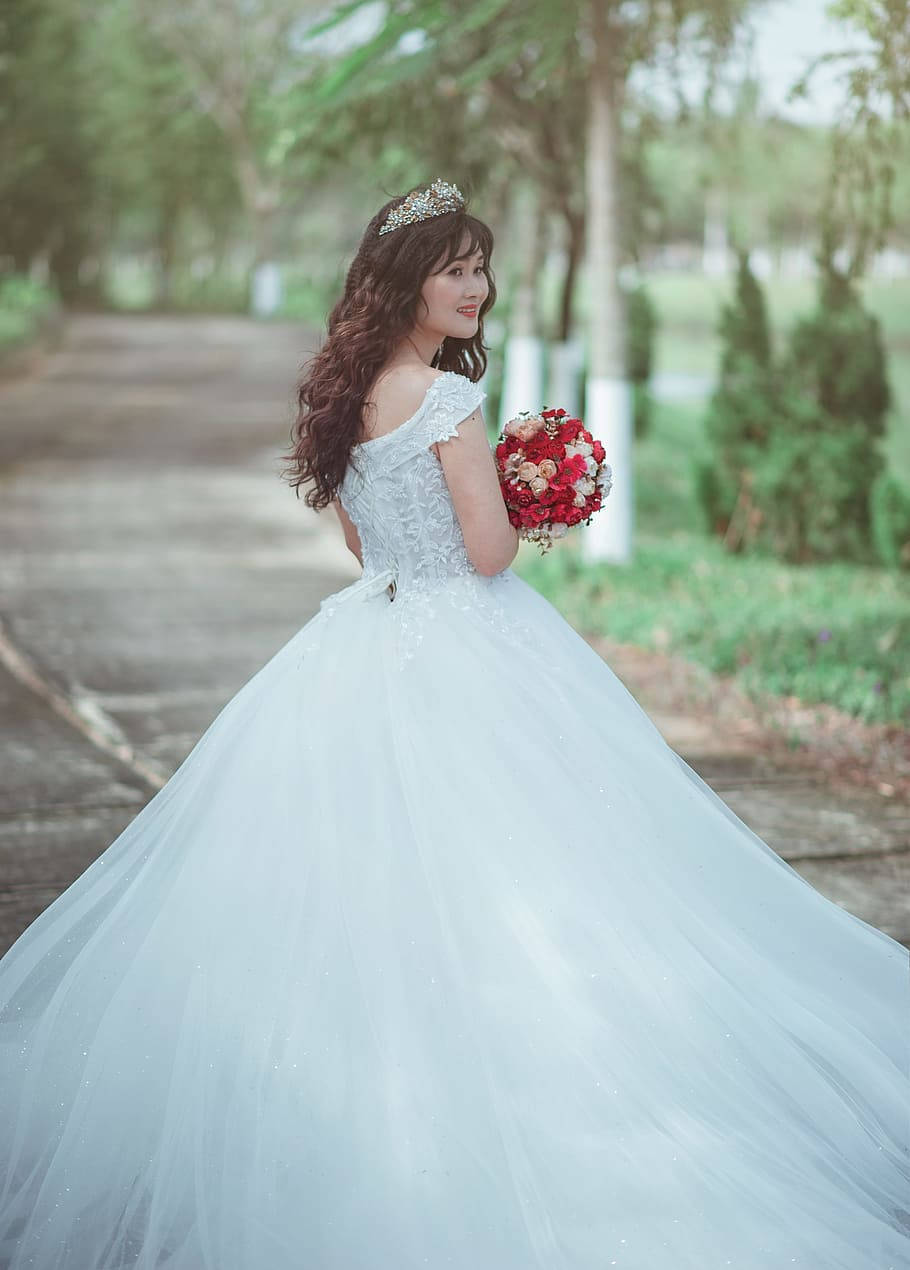 經典婚紗的永恆優雅 壁紙 — 一位身穿閃亮白色婚紗、頭戴皇冠、手持紅色花束的新娘，站在林蔭小徑上，回頭微笑。