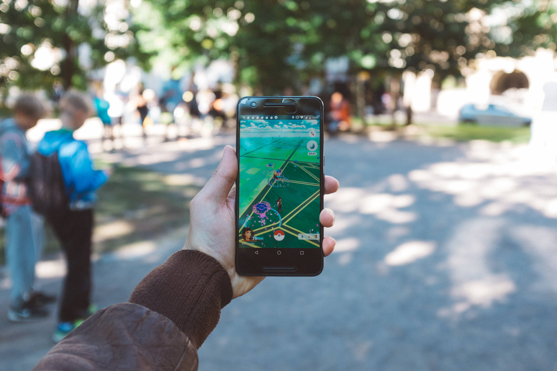 手拿著電話在陽光明媚的公園中玩 Pokémon Go，背景有人和樹木。