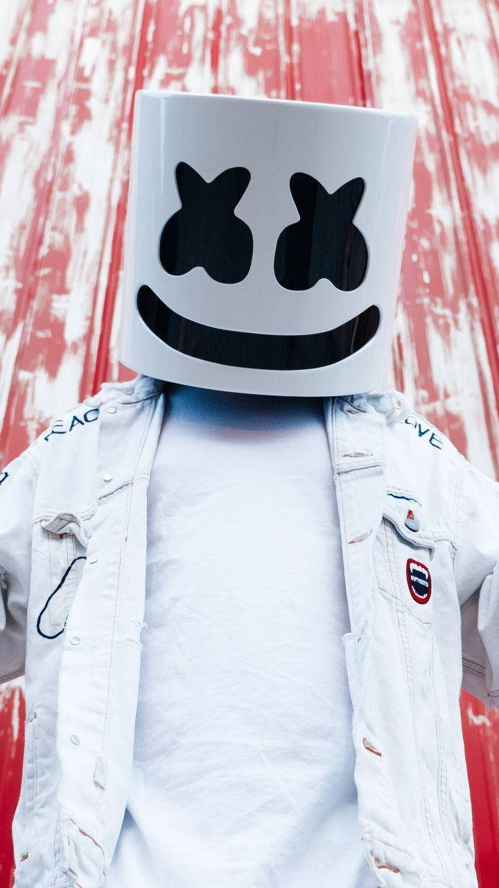 經典微笑 Marshmello iPhone 壁紙 — 一個人戴著白色Marshmello頭盔，站在風化的紅白木牆前。