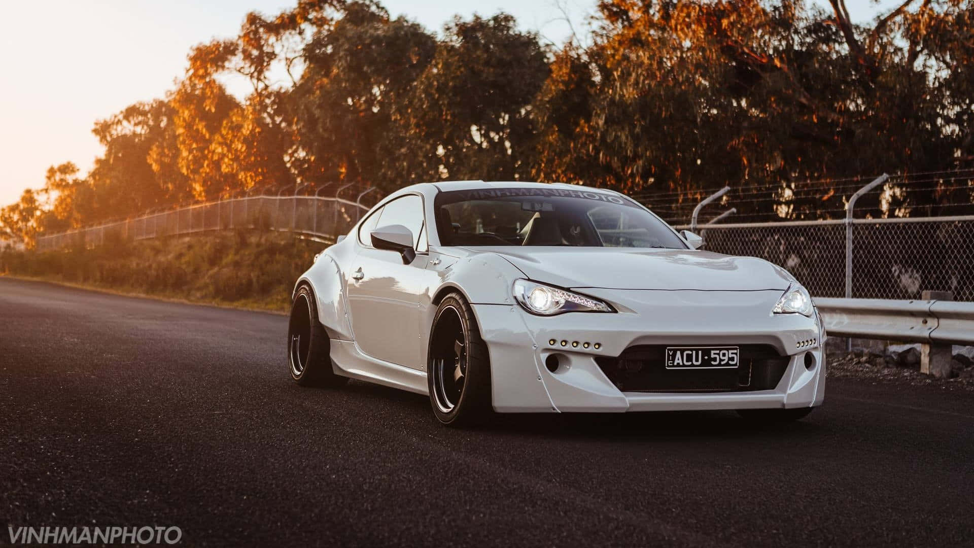 經典敞篷跑車 — Toyota 86 壁紙 — 一架經過改裝的白色流線型跑車，在陽光照耀的瀝青路上滑行，路旁有樹木和圍欄。