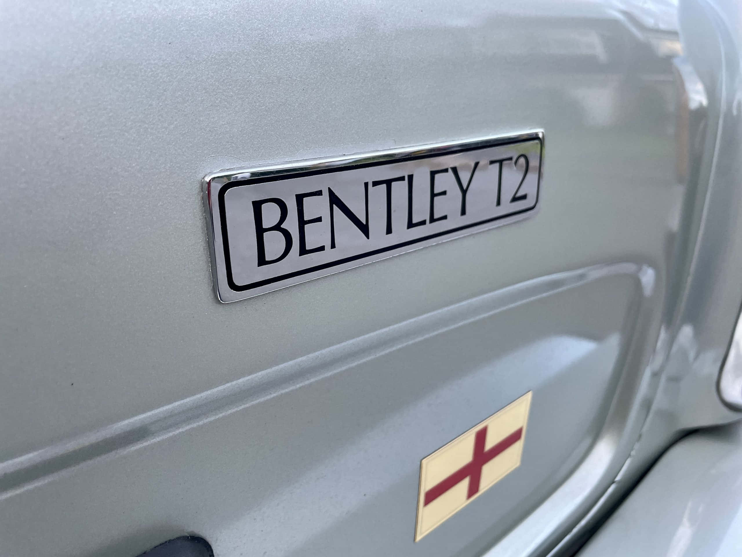 Bentley T2 桌布 壁紙 — 經典汽車擋泥板上帶有紅色十字徽章的銀色 Bentley T2 標誌特寫。