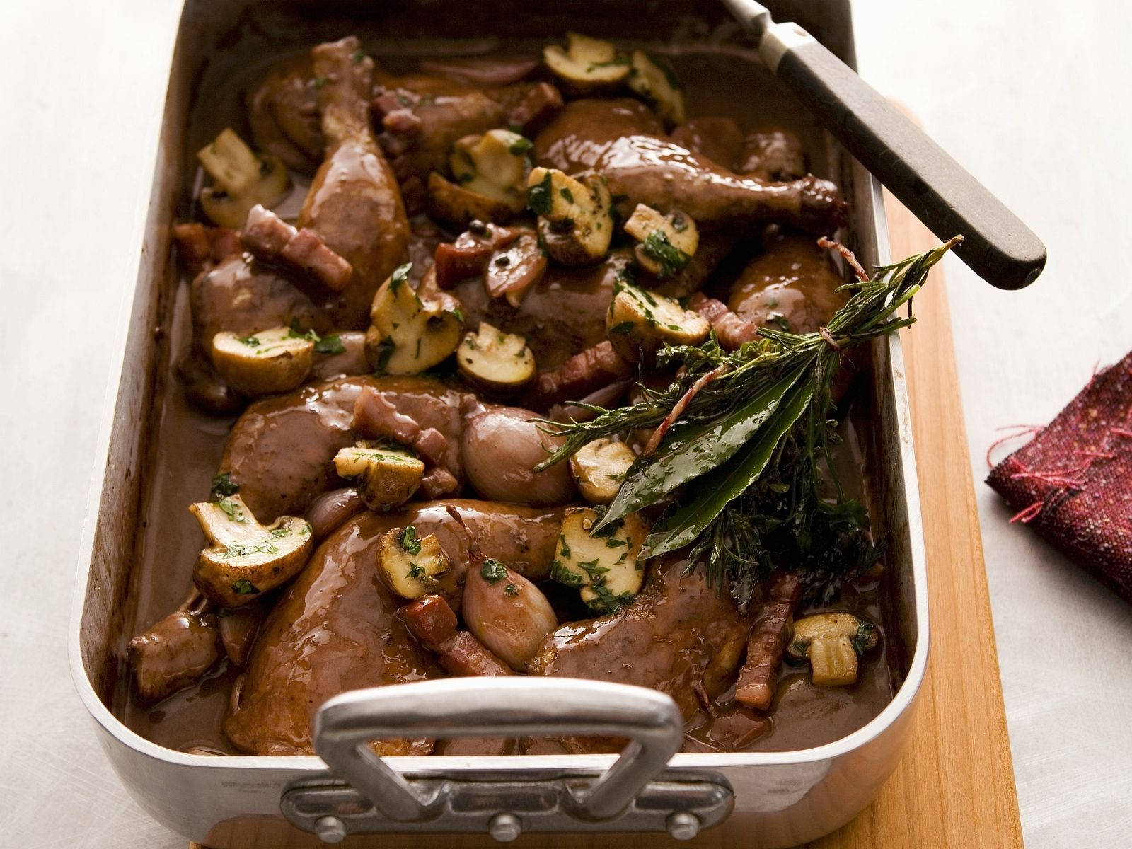 經典法式料理- Coq Au Vin 壁紙 — 濃郁的棕色燉鴨腿、蘑菇和香草在金屬烤盤中慢煮，旁邊有木砧板和紅色餐巾。