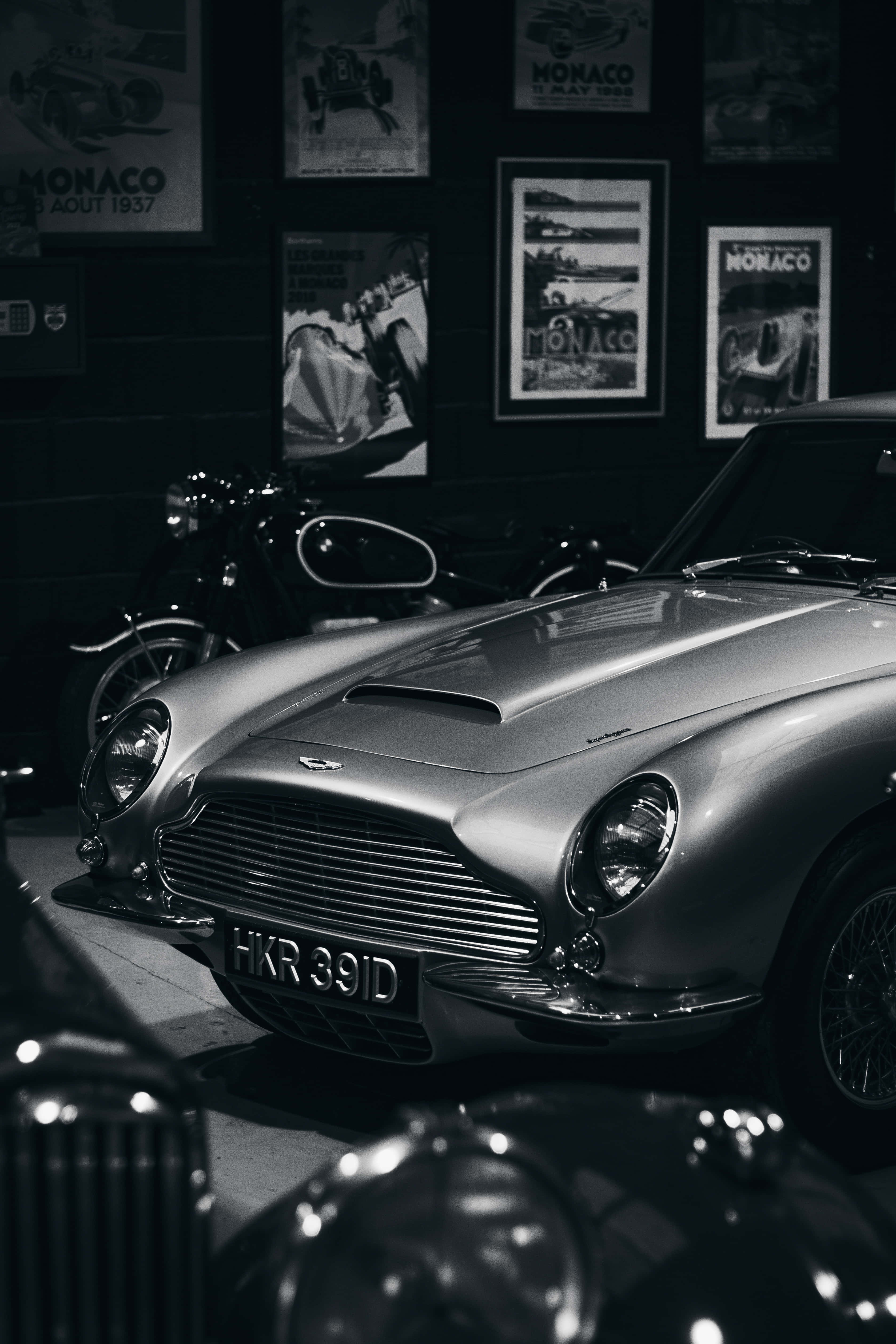 經典法拉利風格 壁紙 — 一輛時尚的銀色經典 Aston Martin DB5 在昏暗燈光下閃耀，位於裝飾有摩納哥賽車海報的復古汽車博物館中。